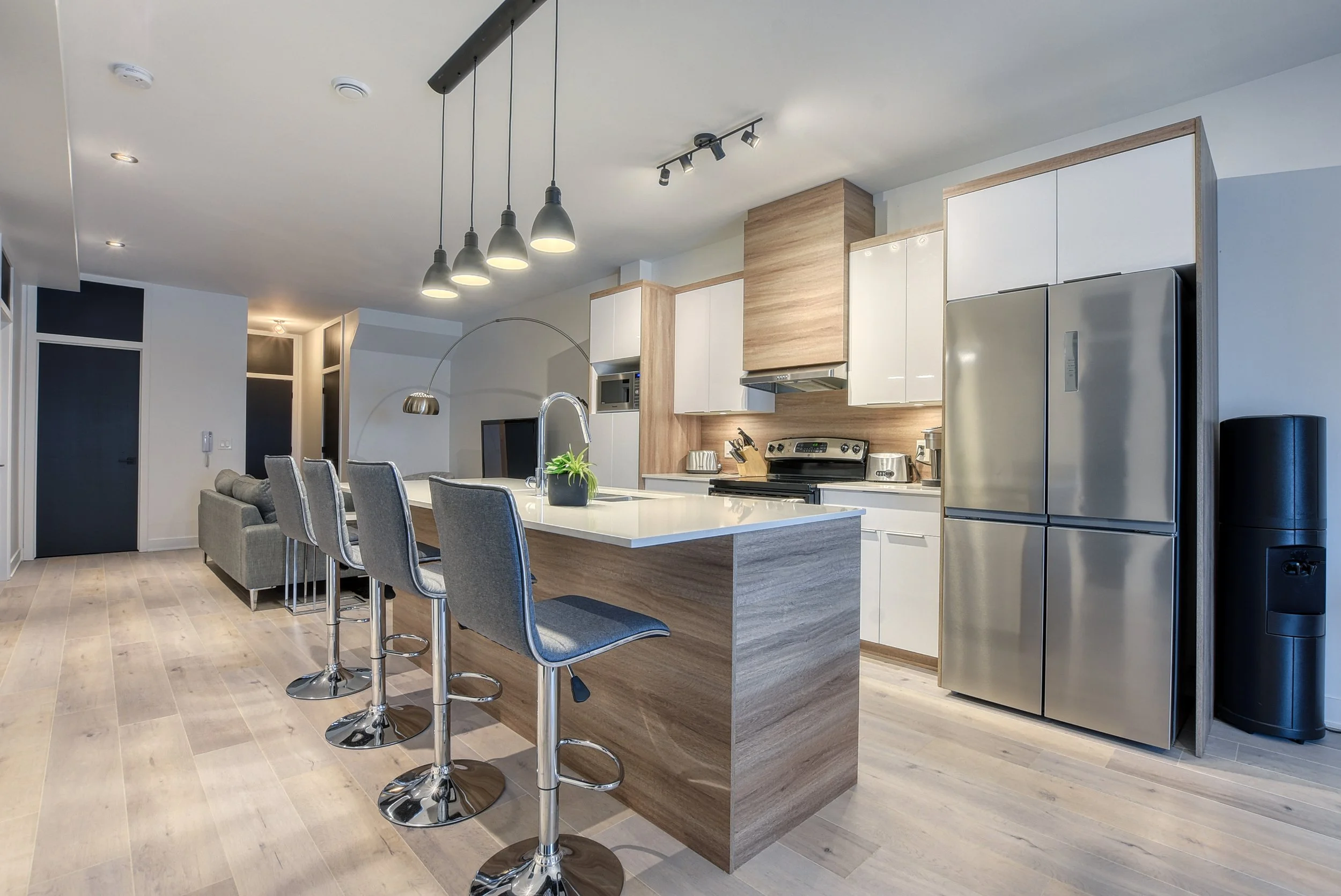 Cuisine moderne avec îlot central, four, réfrigérateur en acier inoxydable, chaises hautes delux, salon avec canapé en arrière-plan, éclairage suspendu et mur en panneaux de bois