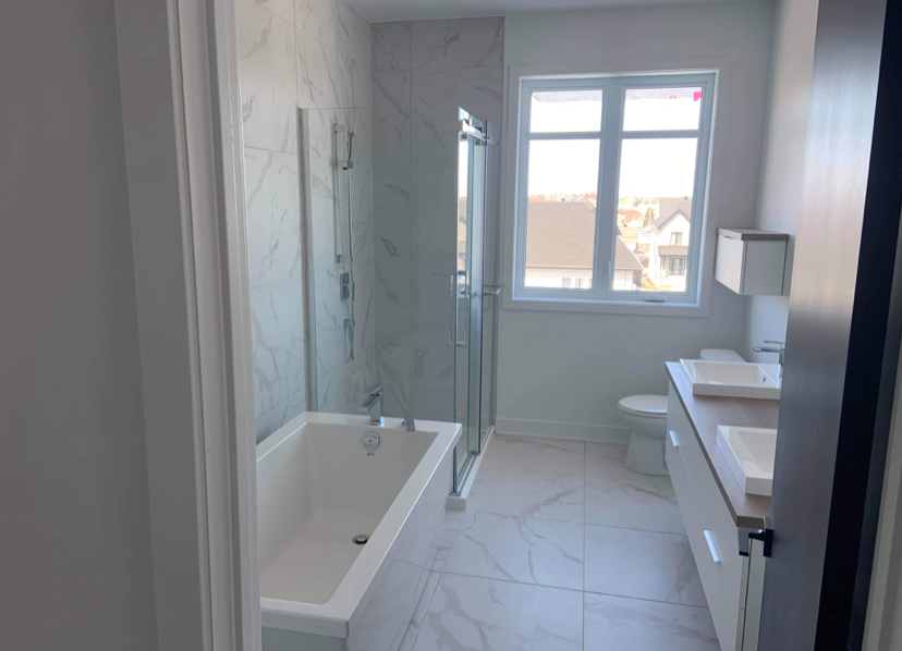 Salle de bain moderne avec une baignoire, une douche en verre, un lavabo sur meuble, une toilette, et une grande fenêtre laissant entrer la lumière naturelle.