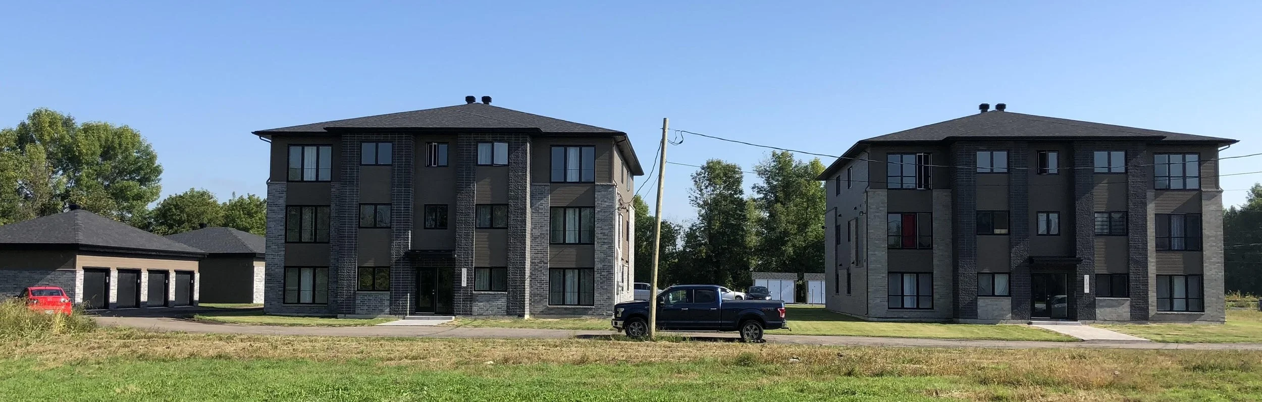 Deux immeubles résidentiels modernes avec de grandes fenêtres, un parking avec des voitures, et un ciel bleu clair.