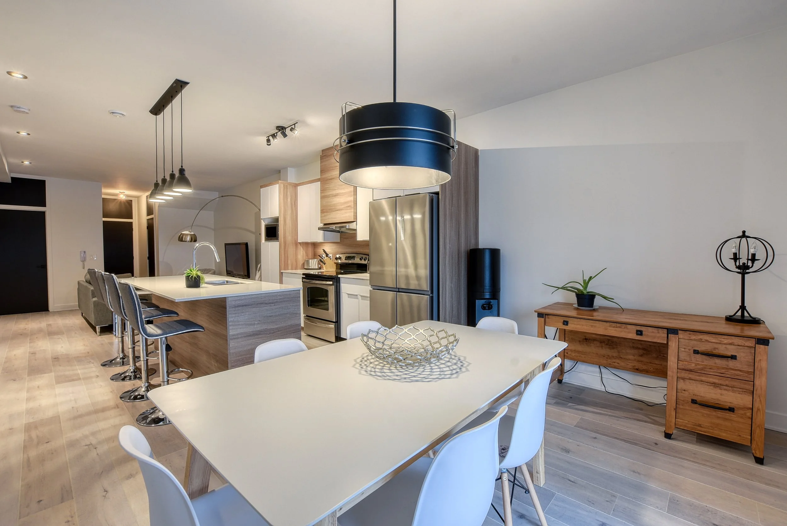 Salon et cuisine modernes avec îlot central, table à manger, mobilier en bois clair, planchers en bois, décor minimaliste avec des plantes et un chandelier