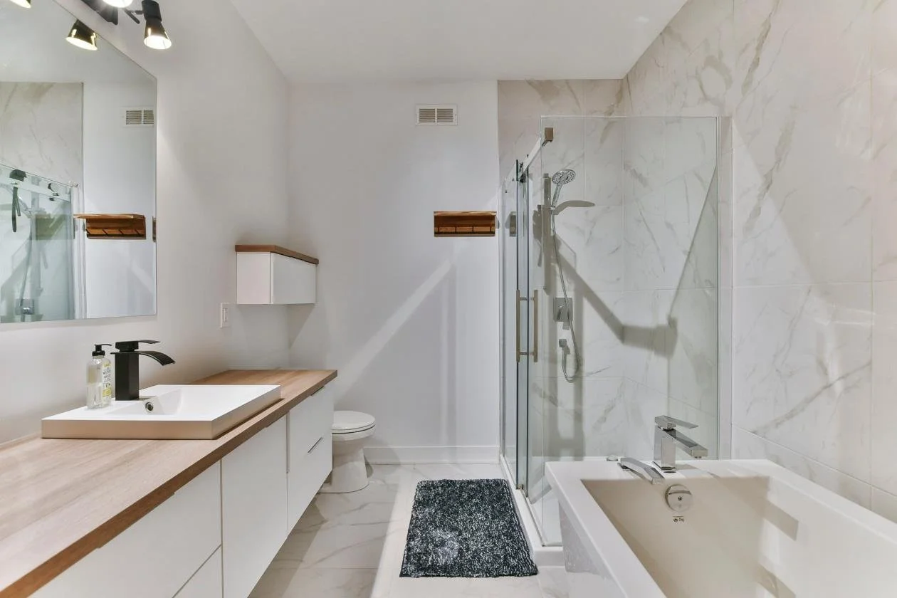 Salle de bain moderne avec douche à l'italienne, baignoire, lavabo sur plan avec miroir et étagères en bois, tapis noir, murs en marbre blanc, et plafond blanc.