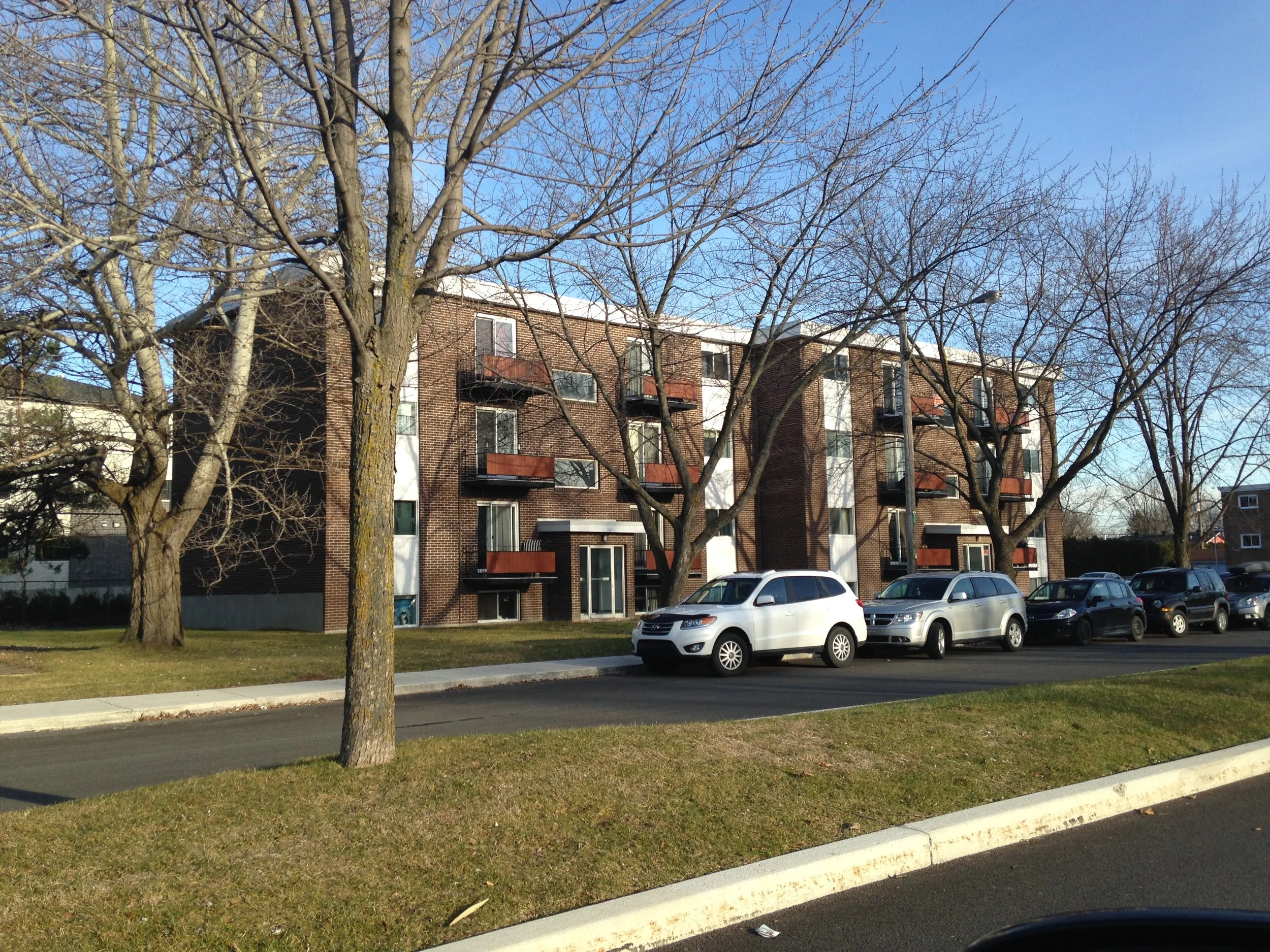 Un immeuble résidentiel avec plusieurs balcons, entouré d'arbres sans feuilles et des voitures garées devant, dans un contexte urbain.