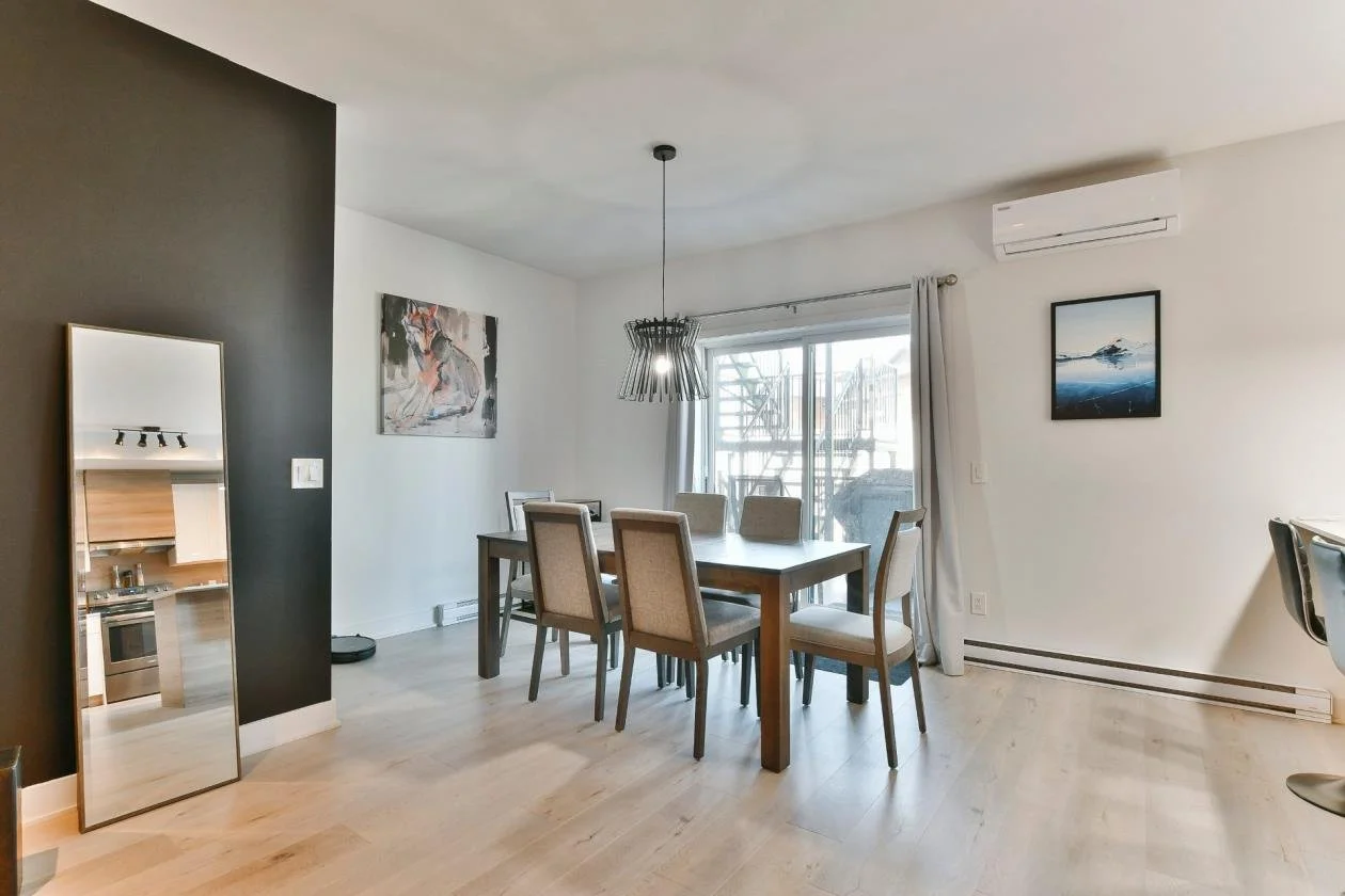 Salle à manger avec une table en bois entourée de six chaises, deux peintures accrochées au mur, une grande fenêtre avec rideau blanc, un miroir réfléchi dans un coin, un climatiseur mural, et une décoration minimaliste.