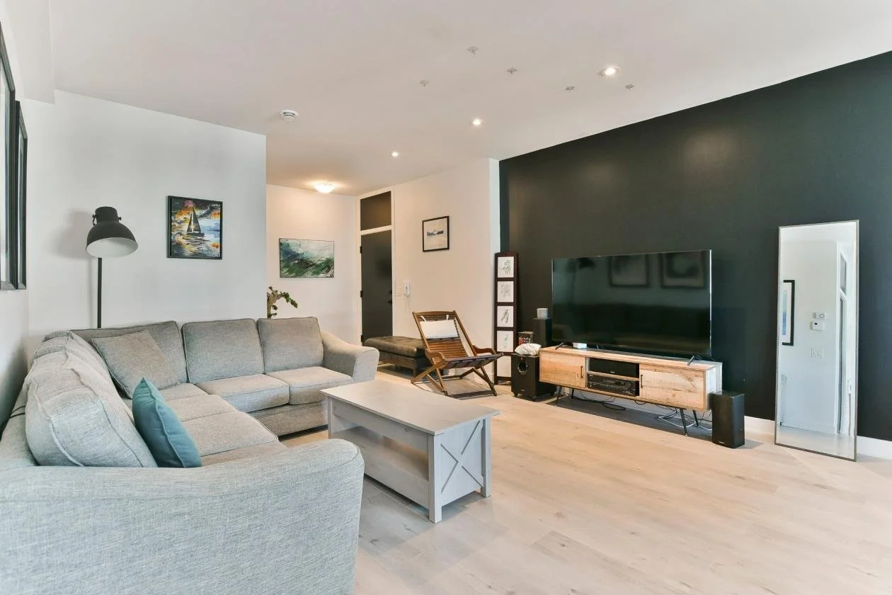 Salon moderne avec canapé gris, télévision à écran plat sur un meuble en bois, mur peint en noir, décoration minimaliste, peinture sur le mur, miroir, chaise en bois, lampe à pied.