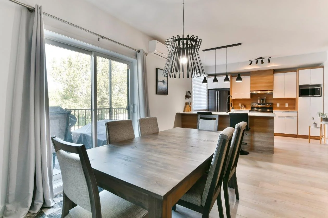 Salle à manger attenante à une cuisine ouverte, avec un grand tableau en bois, chaises rembourrées, porte-fenêtre ouvrant sur un balcon