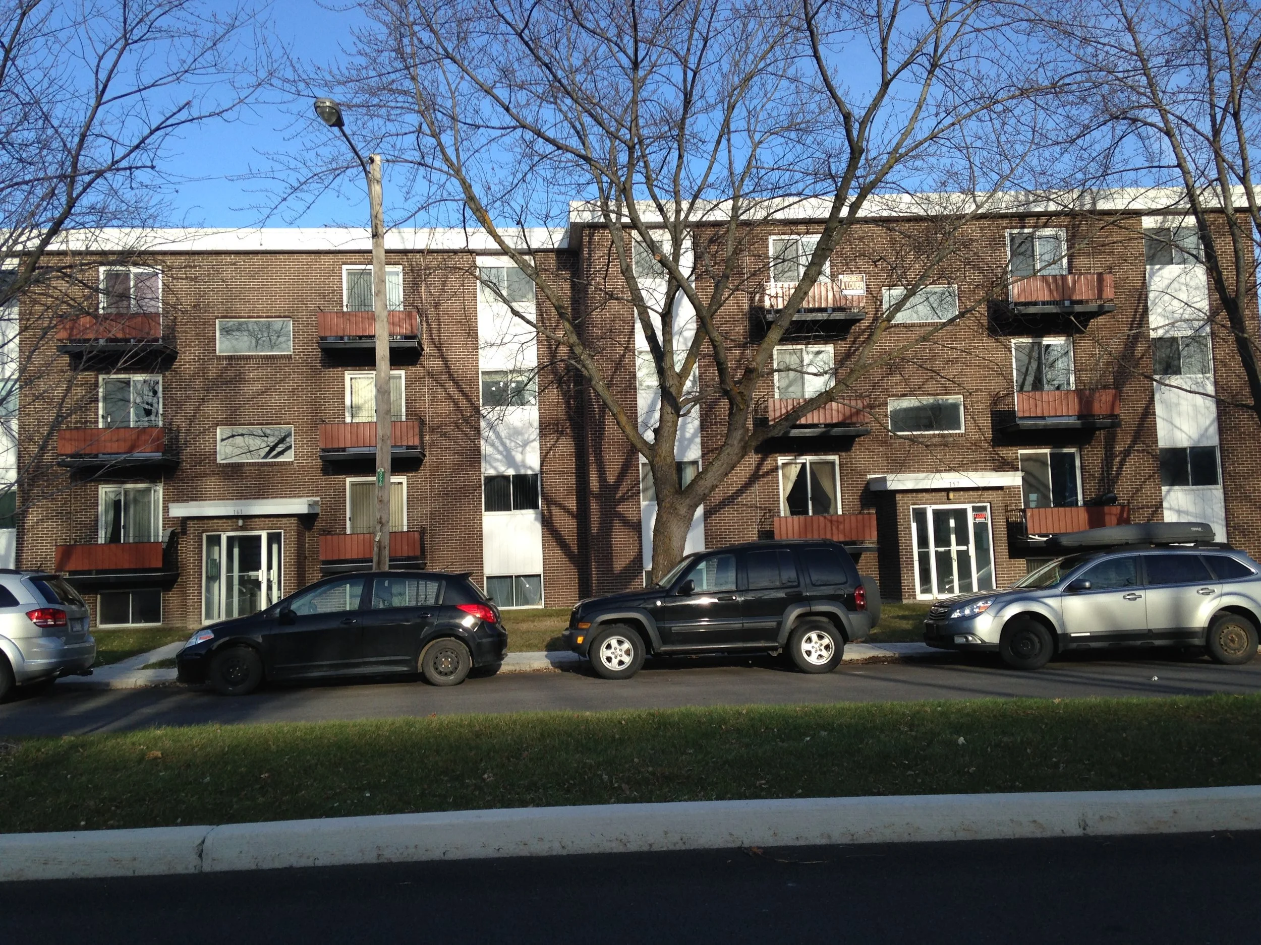 Immeuble résidentiel en brique avec plusieurs balcons, arbres sans feuilles, voitures garées devant, rue en avant-plan, ciel bleu
