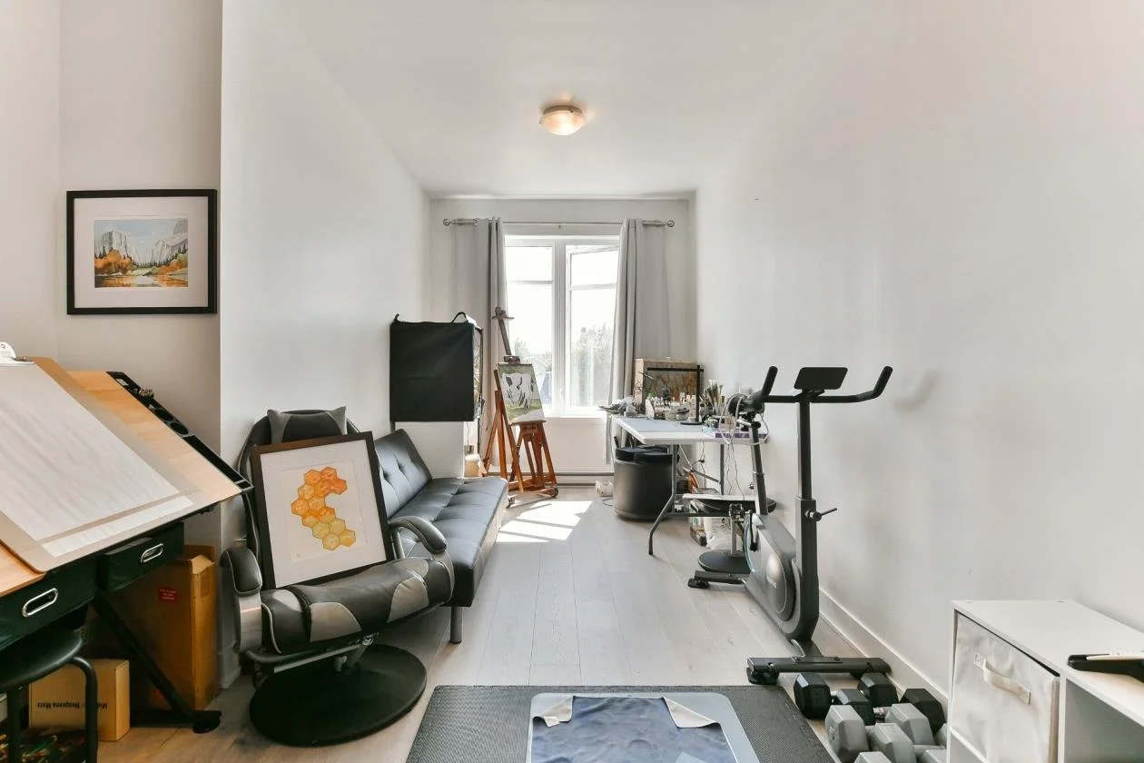 Une pièce d'intérieur avec un canapé noir, un tableau encadré, des appareils de fitness, un bureau avec des objets d'art et une fenêtre laissant entrer la lumière.