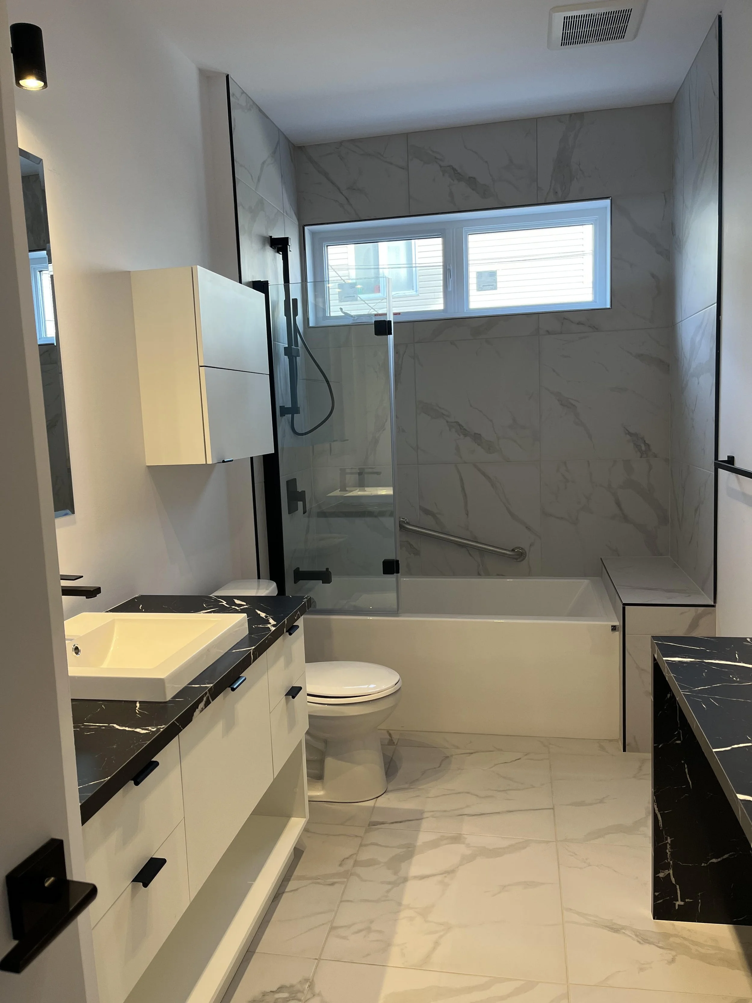 Une salle de bain moderne avec des murs en marbre, un lavabo blanc sur un comptoir noir, un miroir, une toilette, une baignoire avec une paroi en verre et une fenêtre haute permettant la lumière naturelle.