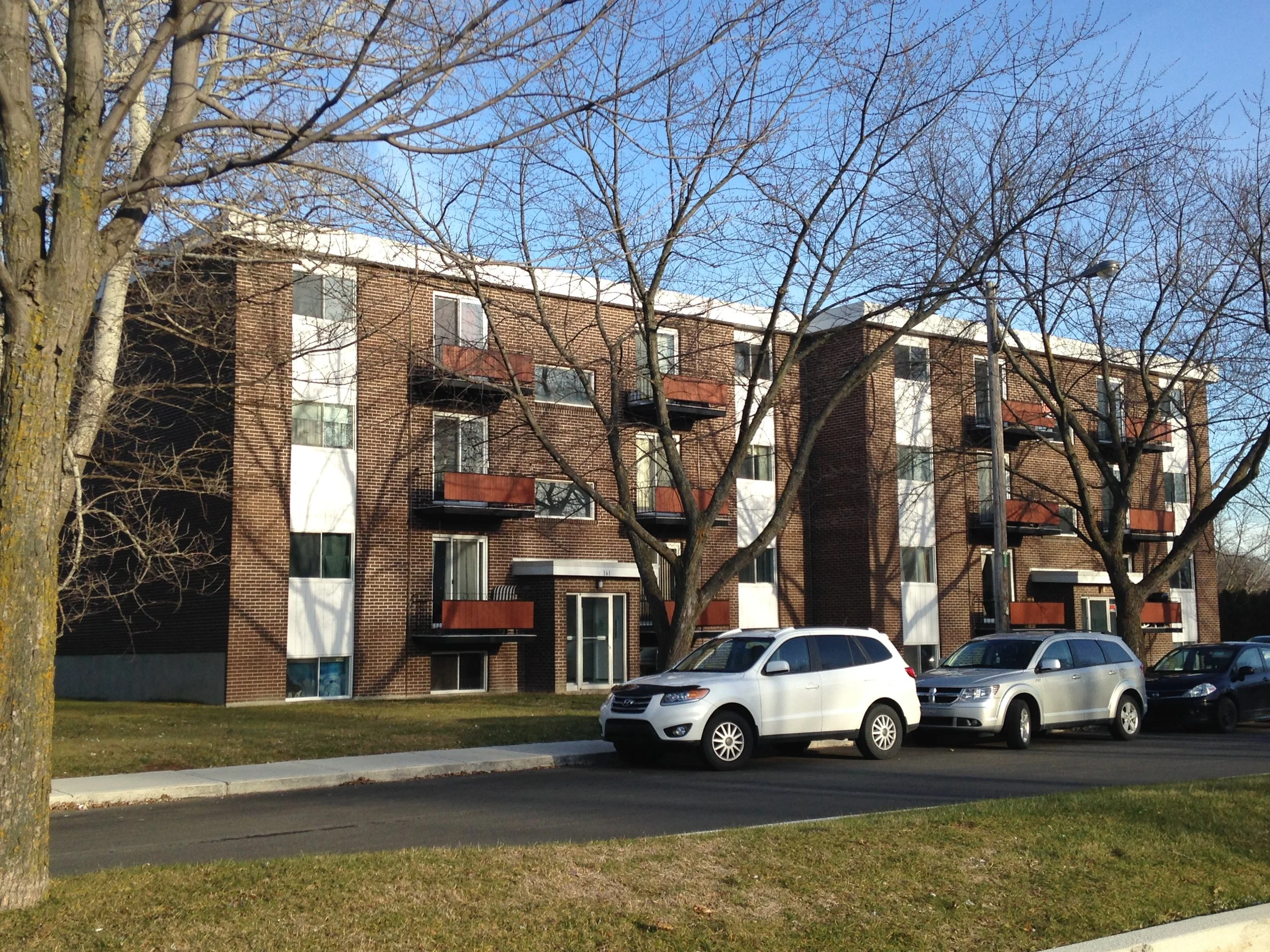 Immeuble résidentiel en briques avec arbres et voitures garées devant, ciel bleu clair.