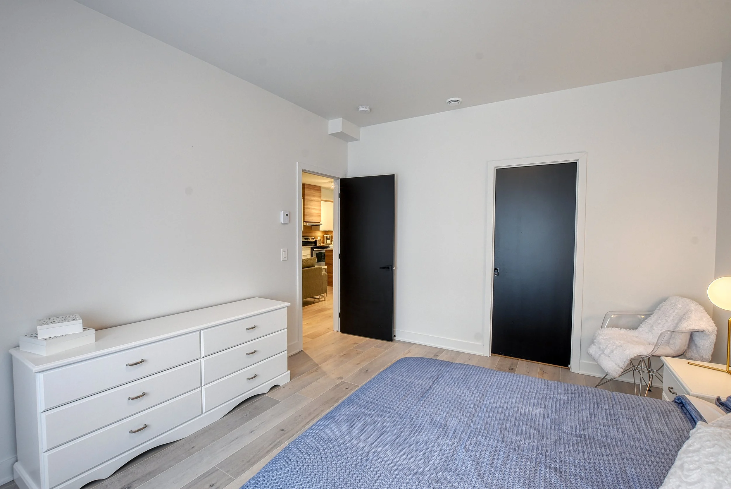 Chambre moderne avec lit, commode blanche, fauteuil blanc avec couverture, porte noire entrouverte, porte noire fermée, et porte aux matériaux naturels. La pièce est lumineuse avec décoration minimaliste.