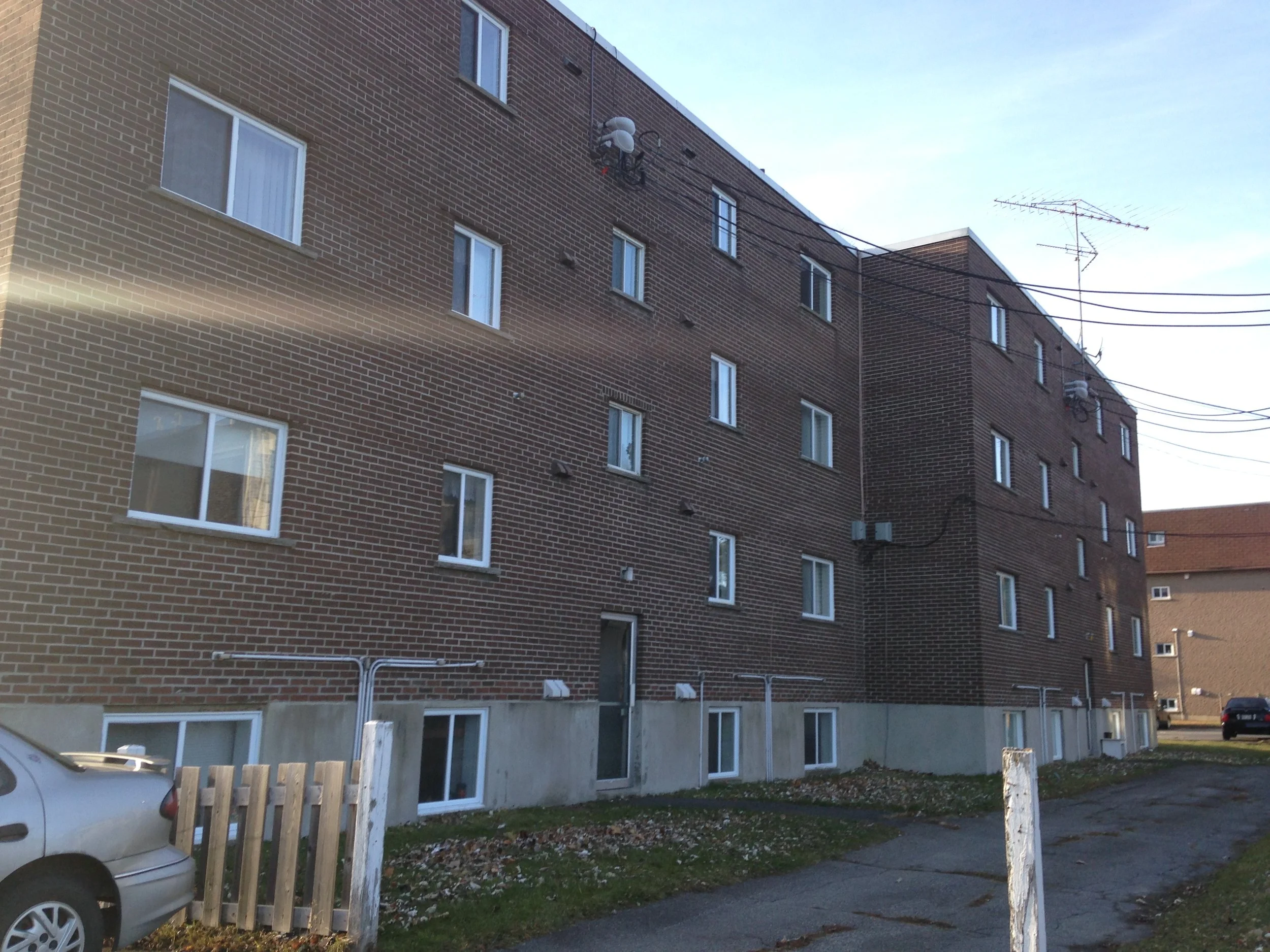 Immeuble résidentiel en briques avec fenêtres, voitures stationnées devant, ciel clair, en zone urbaine.