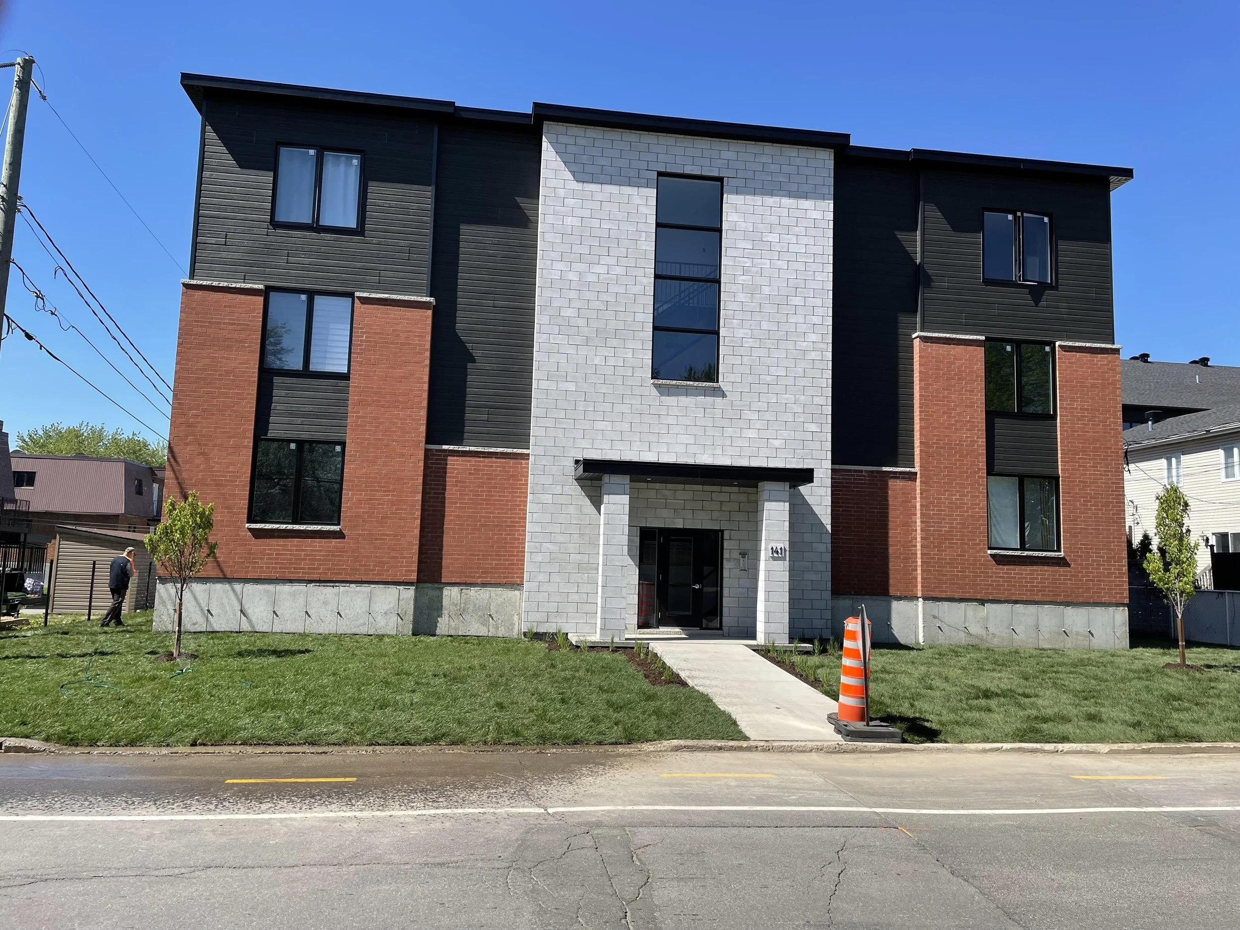Immeuble résidentiel moderne avec façade en briques rouges, panneaux noirs et mur en briques grises, entrée principale sous un petit porche, deux jeunes arbres devant, un cône orange à proximité de l'entrée, ciel bleu clair, route en premier plan.