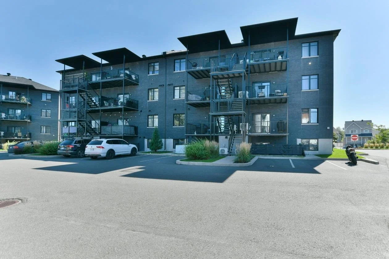 Immeuble résidentiel avec plusieurs étages, escaliers extérieurs et un parking devant. Le bâtiment est en briques foncées et entouré de végétation.