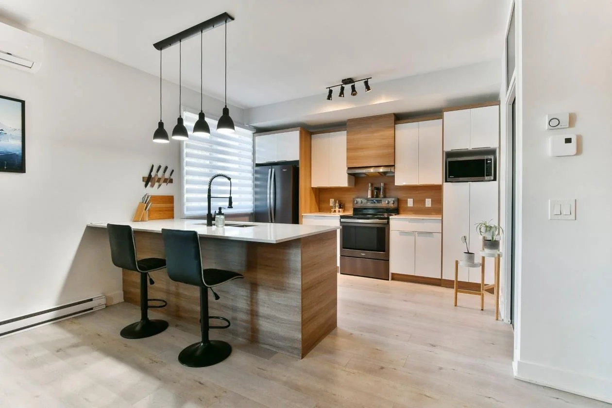 Cuisine moderne avec îlot en bois et tabourets noirs, éclairage suspendu, électroménagers en acier inoxydable, fenêtre avec stores vénitiens, décoration minimaliste, plancher en bois clair.