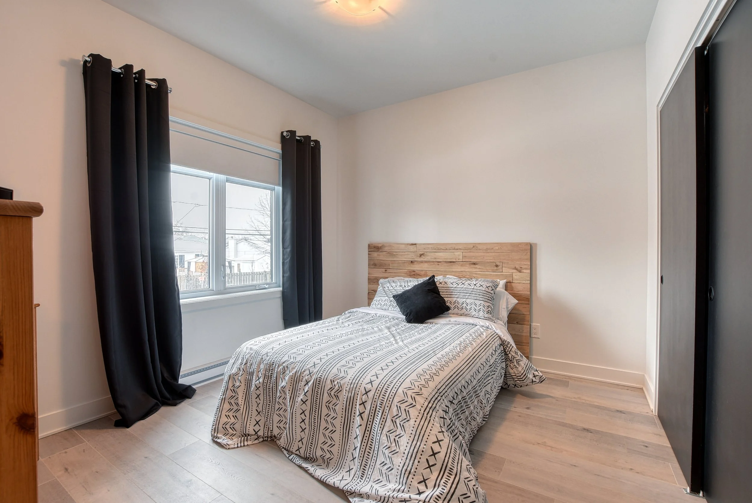 Chambre avec un lit, une fenêtre avec des rideaux noirs, un mur blanc et un sol en bois clair.