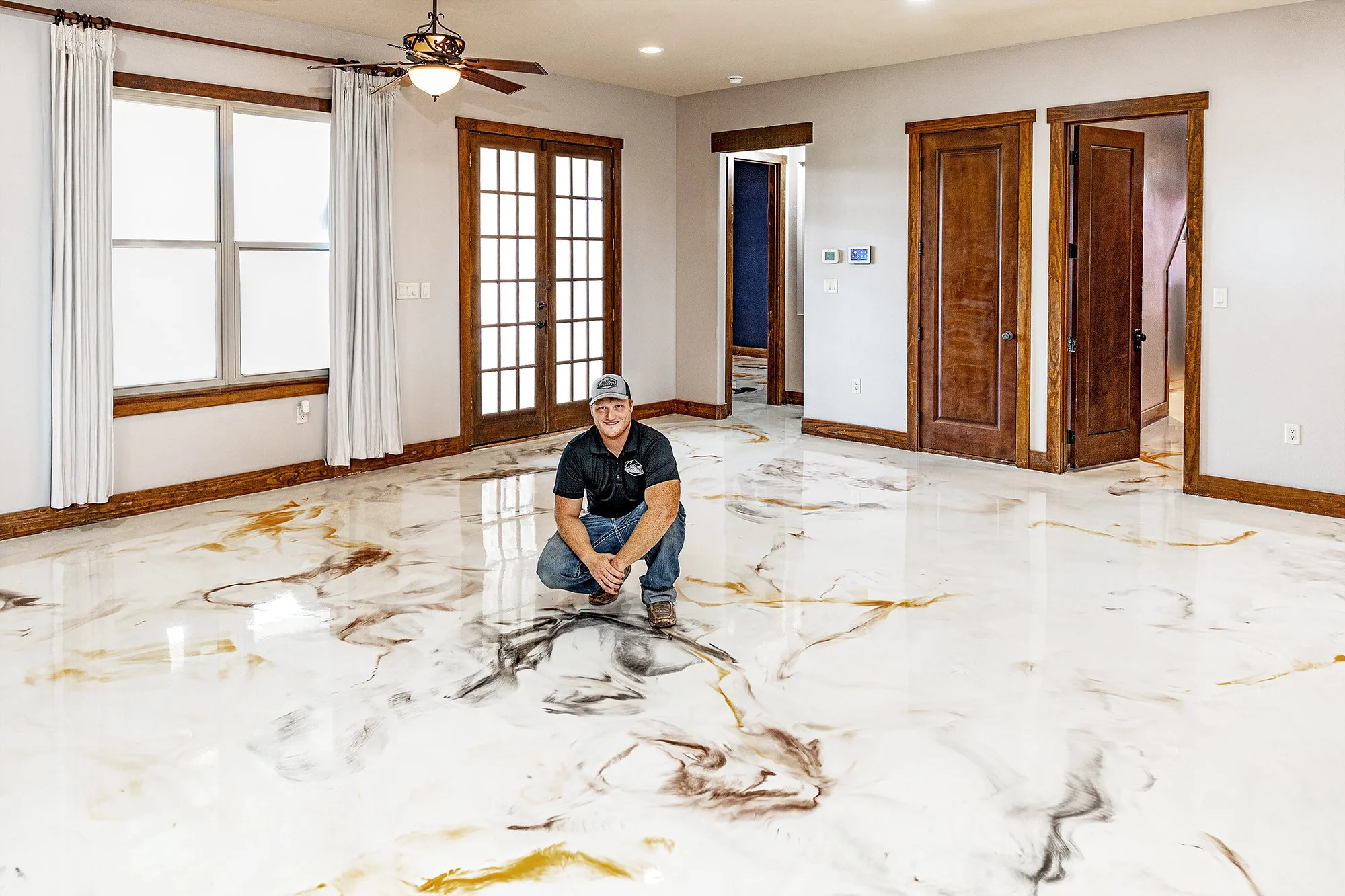 Man crouching on a marble floor in a living room with white walls, wooden trim, large windows with white curtains, and a ceiling fan.