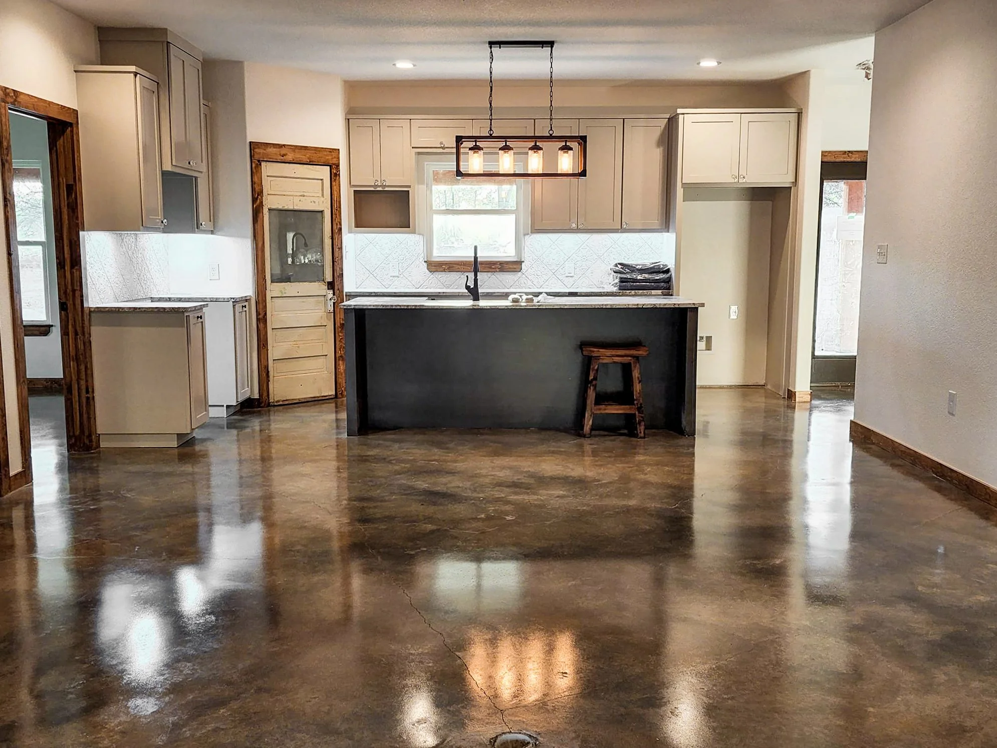 Interior of a kitchen with wooden floors, beige cabinets, and a black kitchen island with a stool. There is a window above the sink, and a light fixture hanging above the island. The walls are painted light, and a door with a glass panel is on the left. The room looks modern and minimalistic.