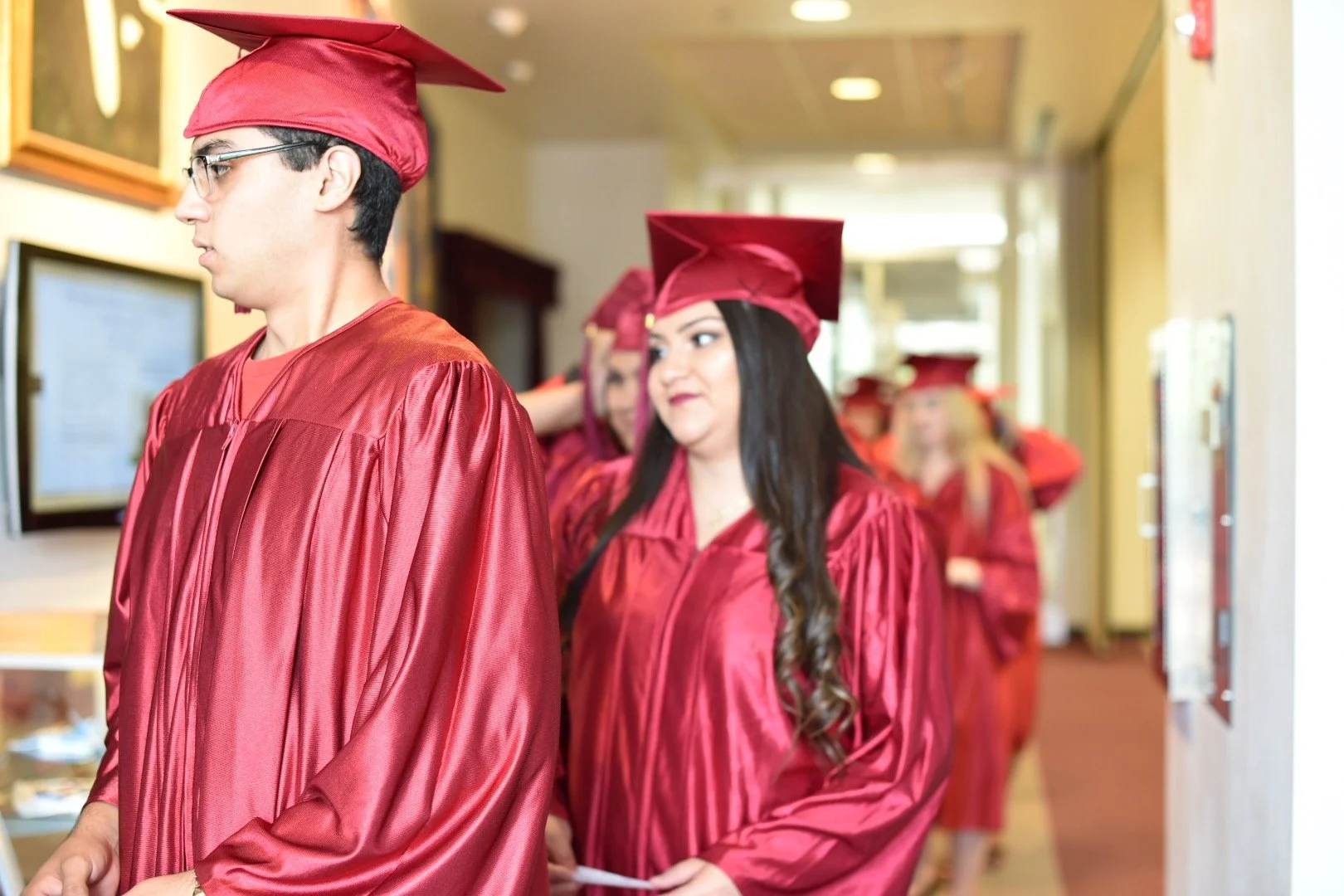 Graduates in red caps and gowns standing in line indoors, preparing for a ceremony.