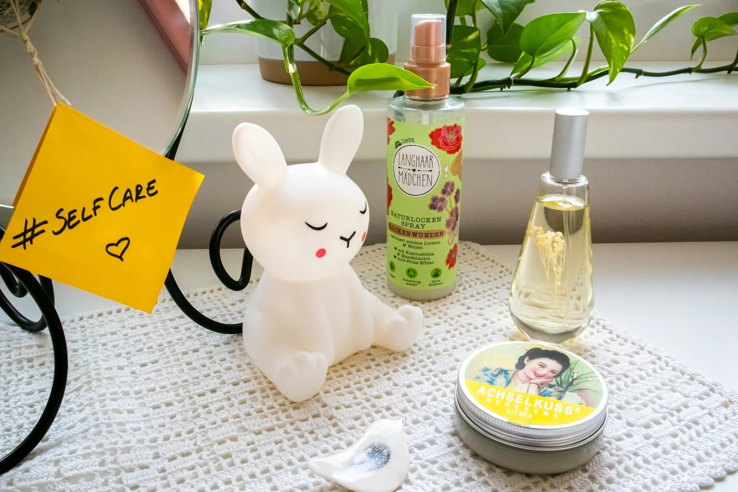 A white bunny-shaped diffuser with closed eyes, surrounded by a spray bottle labeled 'Naturlack Schnuzenwunder,' a glass bottle of liquid with a floral scent, a small tin can with a woman’s photo and text 'Achselkusss Deo Creme,' a heart-shaped dish, and a yellow sticky note with '#SelfCare' written on it, all on a white crocheted cloth in front of a window with green plants.
