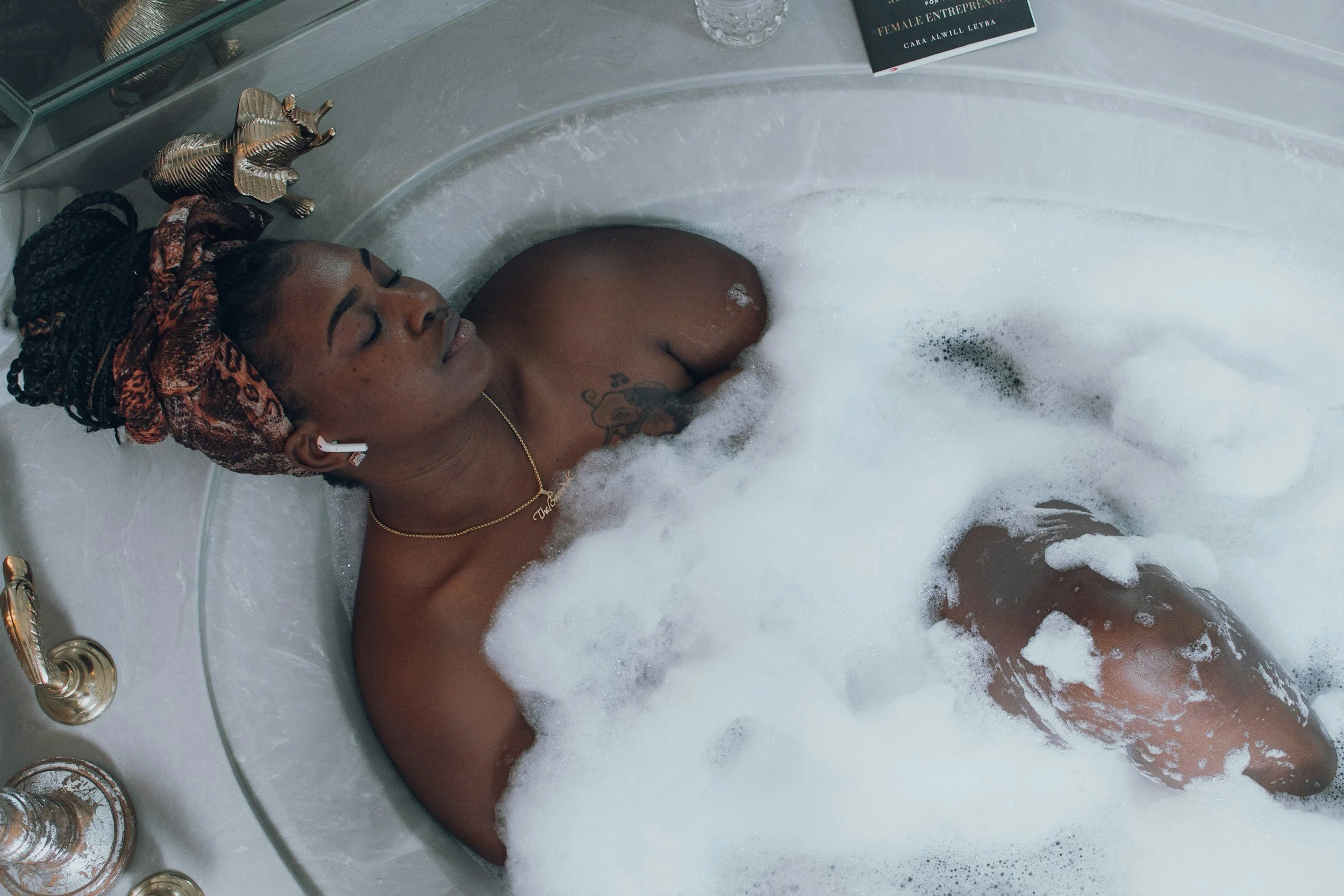 A woman with dreadlocks and tattoos relaxing in a bubble bath with her eyes closed, wearing wireless earbuds and jewelry.