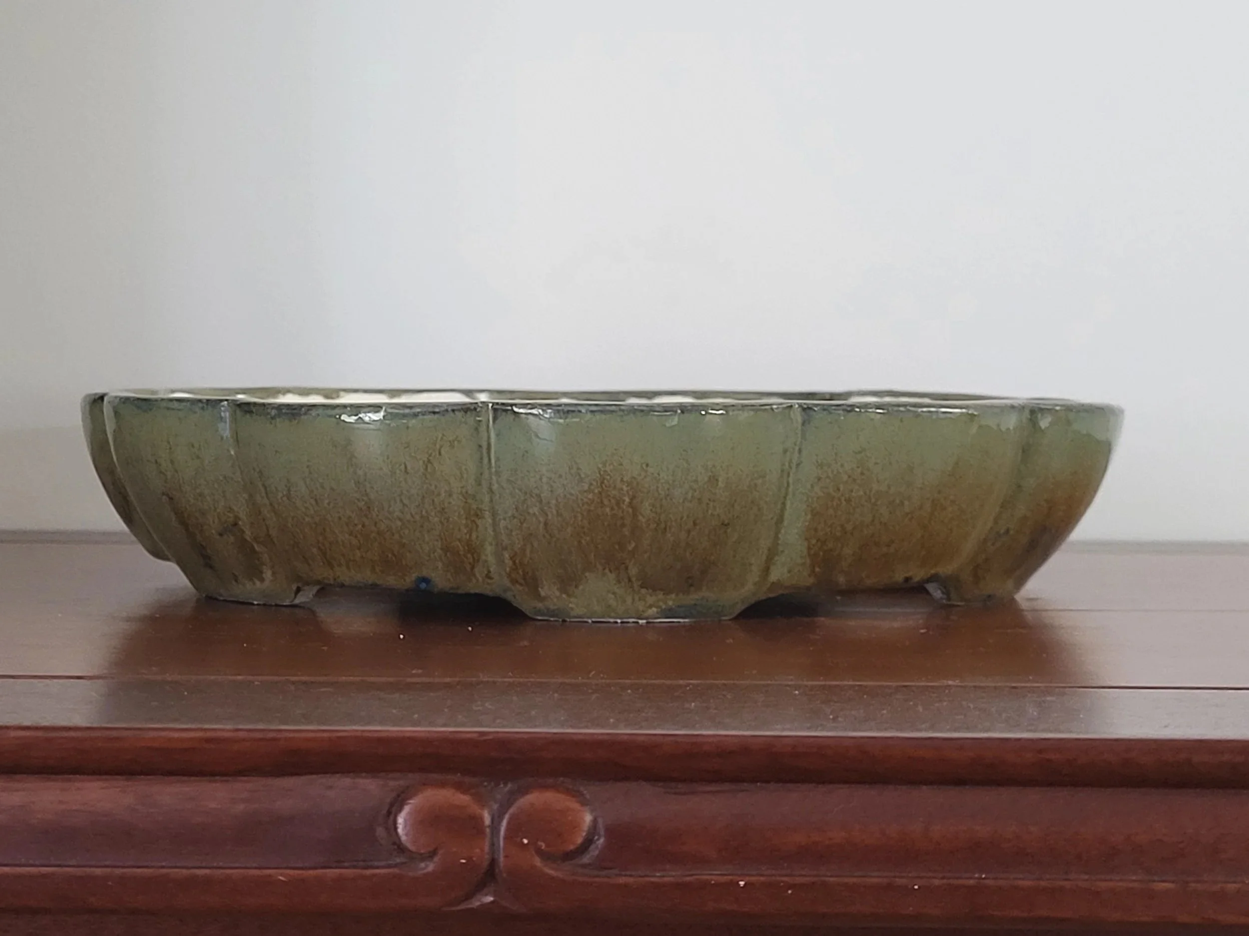 A shallow ceramic bowl with a greenish-brown glaze sitting on a wooden surface against a plain white background.
