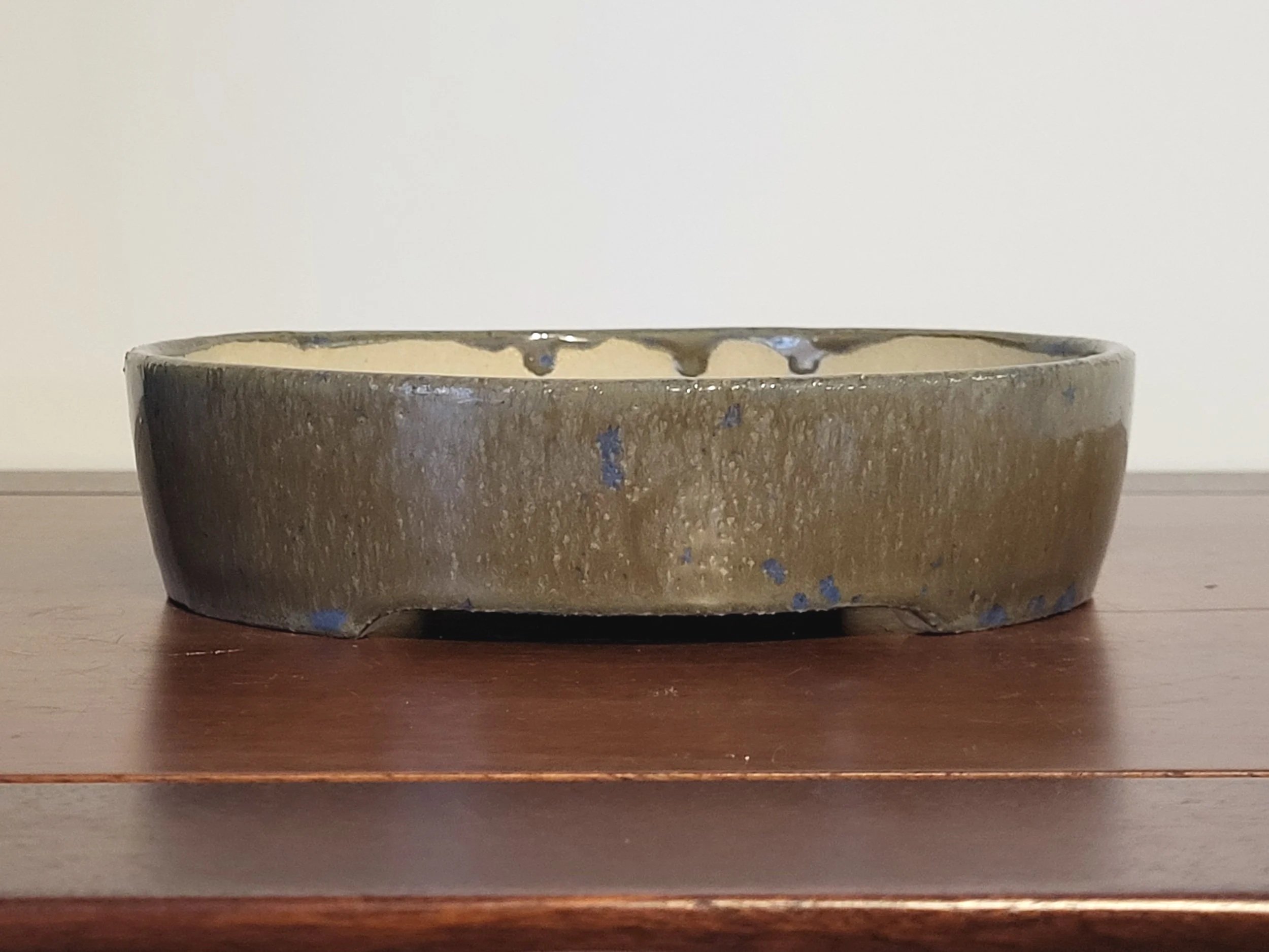 A round, shallow ceramic bowl with a textured, uneven surface, glazed in an earthy brown tone with blue speckles, resting on a wooden surface.