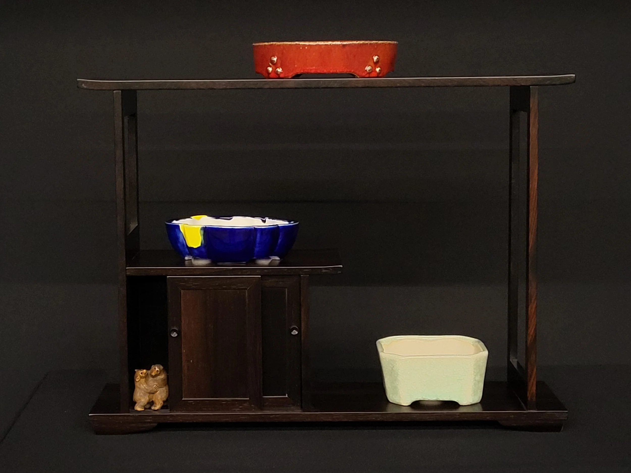 Display of three decorative bowls on a multi-level wooden stand against a black background, with a small lion figurine on the bottom left.