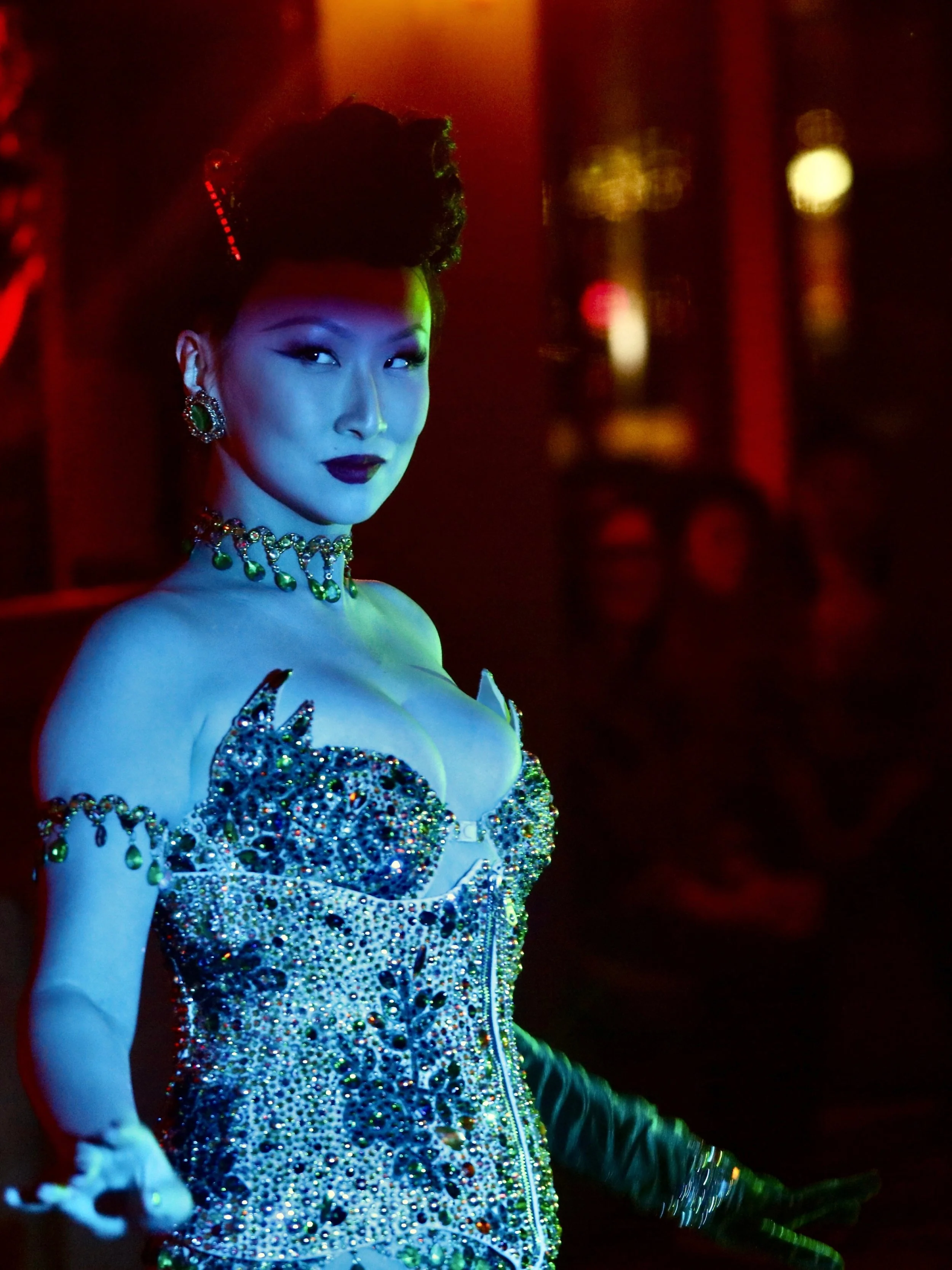 A woman in a glittering, rhinestone-studded dress and jewelry, with dark lipstick, in a nightclub with colorful lighting.