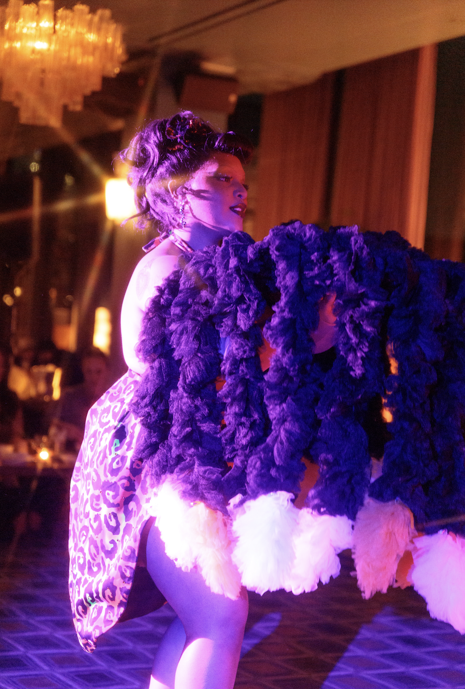 A performer dancing in a nightclub or party setting, dressed in a vibrant animal-print outfit with large feathered accessories, illuminated by colorful stage lighting.
