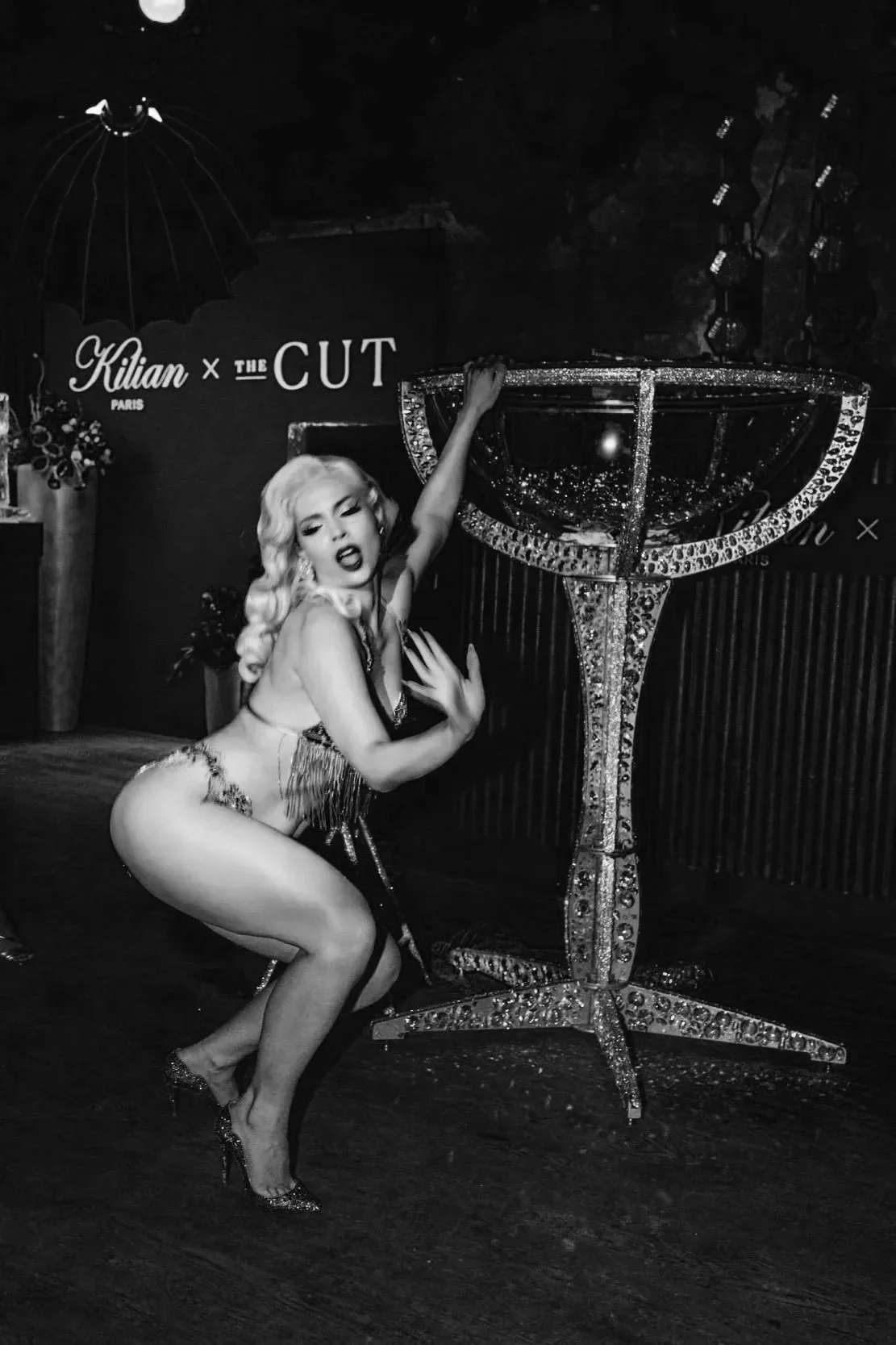 A woman in a glamorous outfit posing next to a large decorated cocktail glass prop at an event or performance.