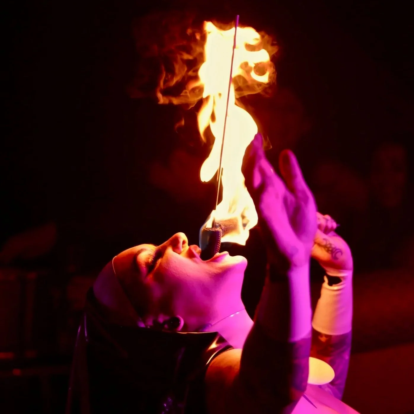 A person with purple lighting on their face and tattoos on their arms is performing a fire-eating act, holding a lit torch close to their mouth with fire engulfing the torch.