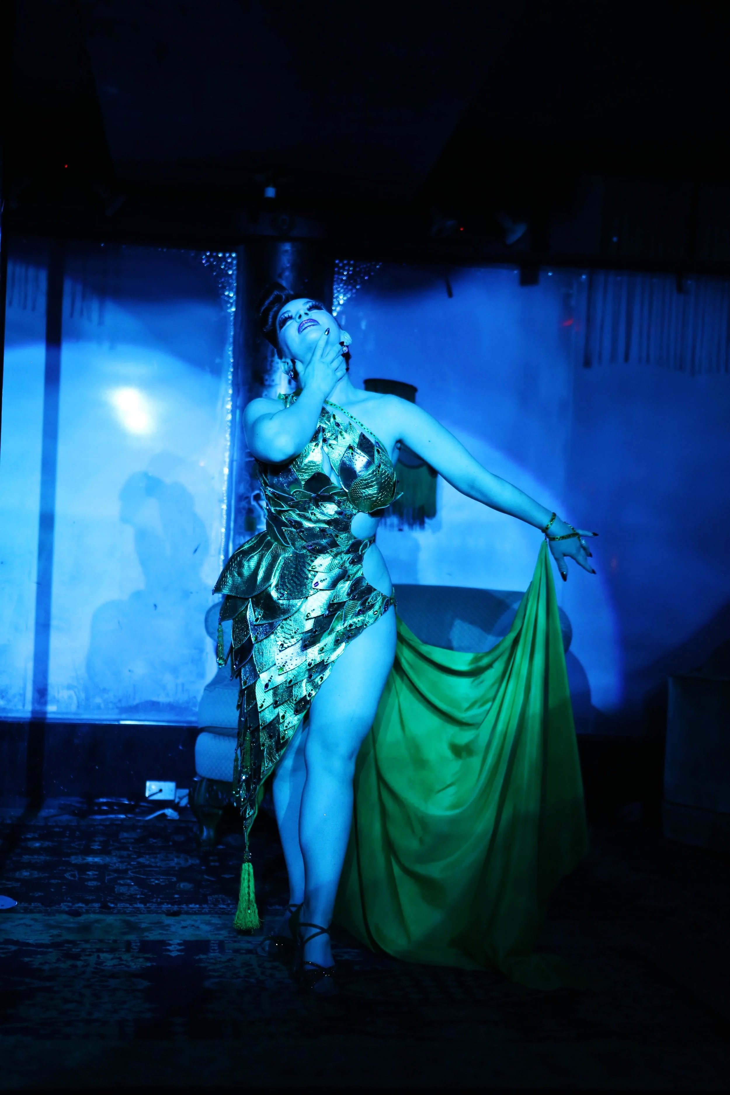 A performer dressed in a gold and black costume with a green fabric, standing on stage under blue lighting, striking a dramatic pose with one hand on her neck and the other holding the fabric.