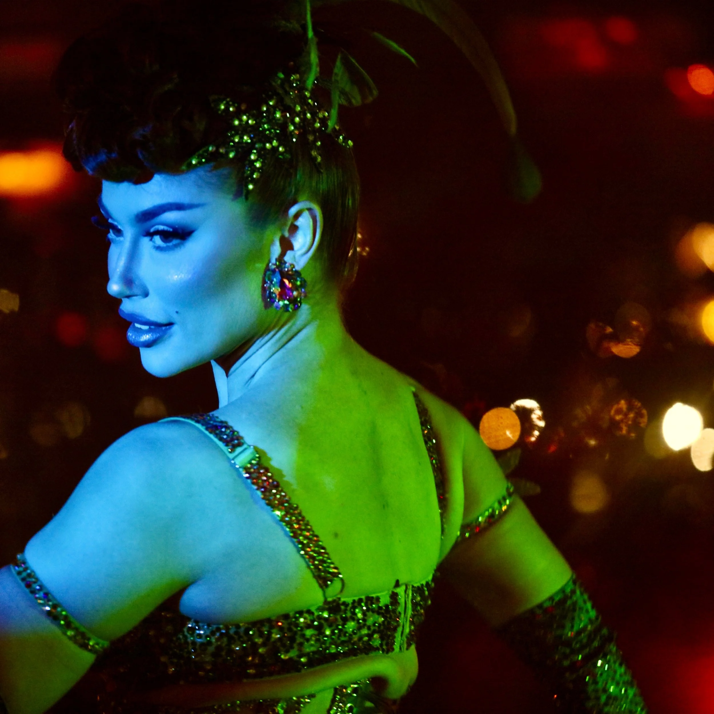 A woman dressed in elaborate, colorful costume with glittering details, posed in a dimly lit setting with colorful bokeh, appearing to be dancing or performing.