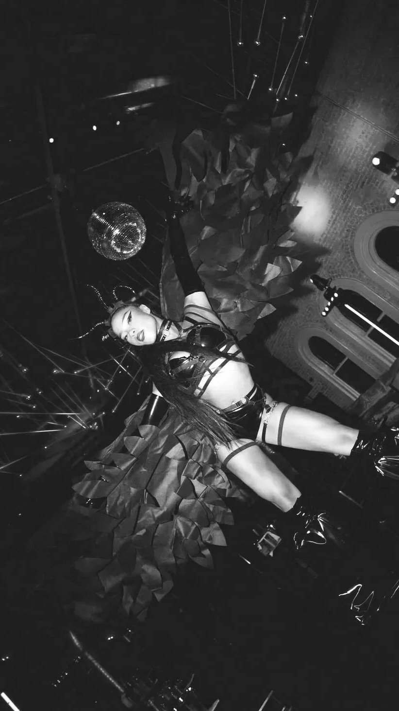 A stilts performer with dark angel wings revealing black leather outfit, stockings, and boots posing on a decorated stage with a disco ball and floral arrangements, illuminated by stage lighting, in a nightclub or performance venue.