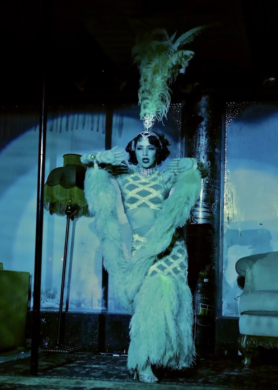 A performer dressed in a glamorous showgirl costume with feathers, a sequined gown, and a feathered headpiece on stage.