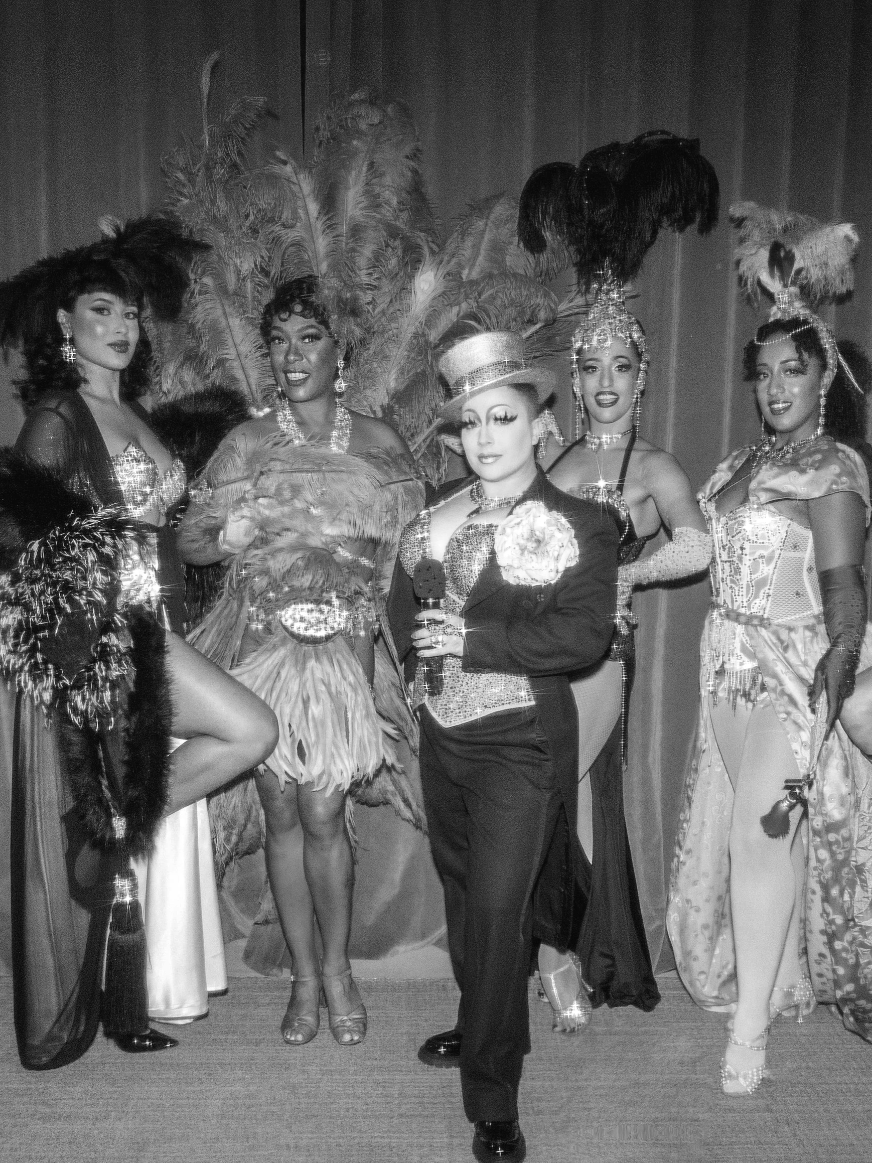 Black and white photograph of women dressed in showgirl costumes with feathered headpieces and elaborate costumes, posing together on stage.