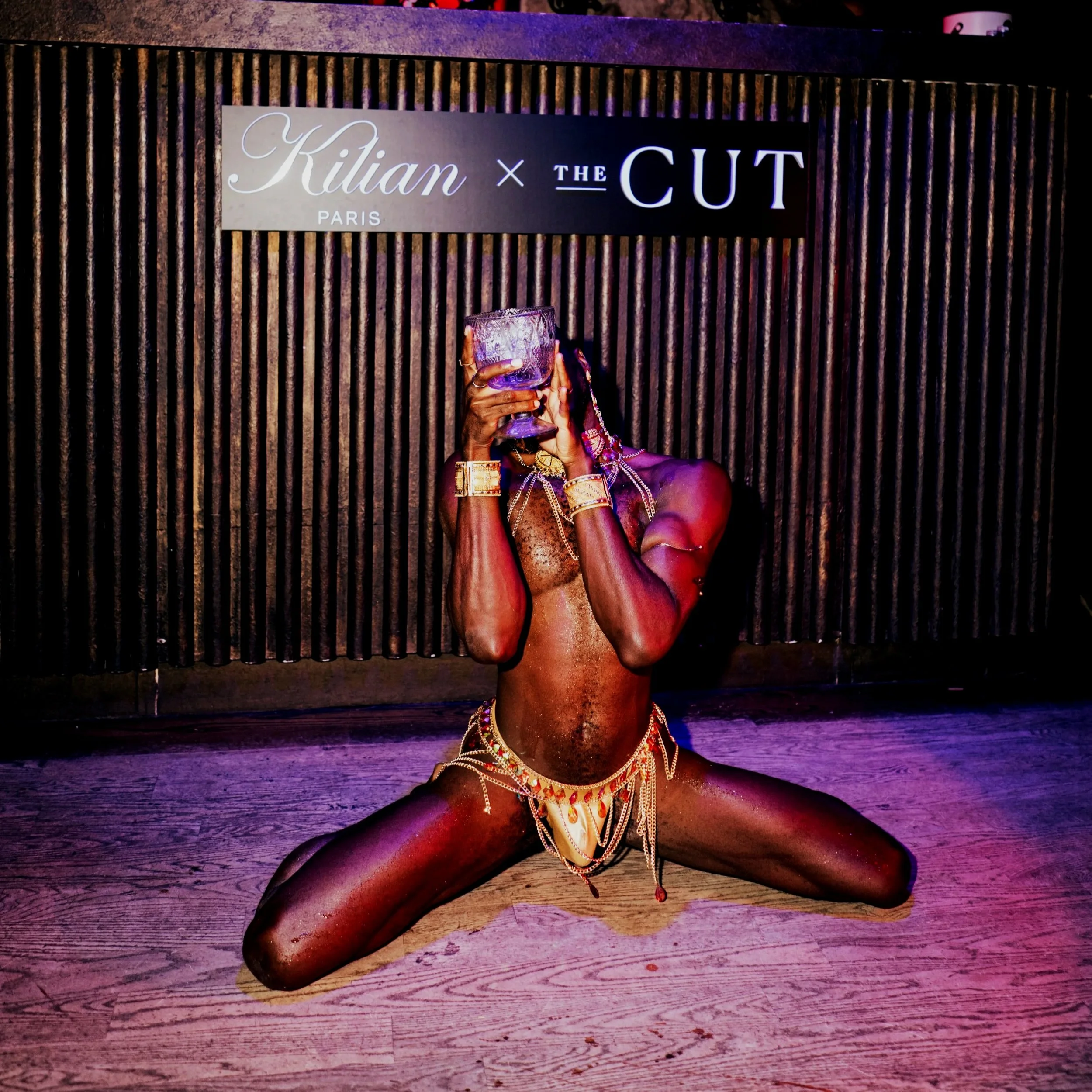 A performer kneeling on the floor with legs spread wide, holding a glass up to their face, in front of a wooden backdrop with a sign that reads 'Kilian × THE CUT'.
