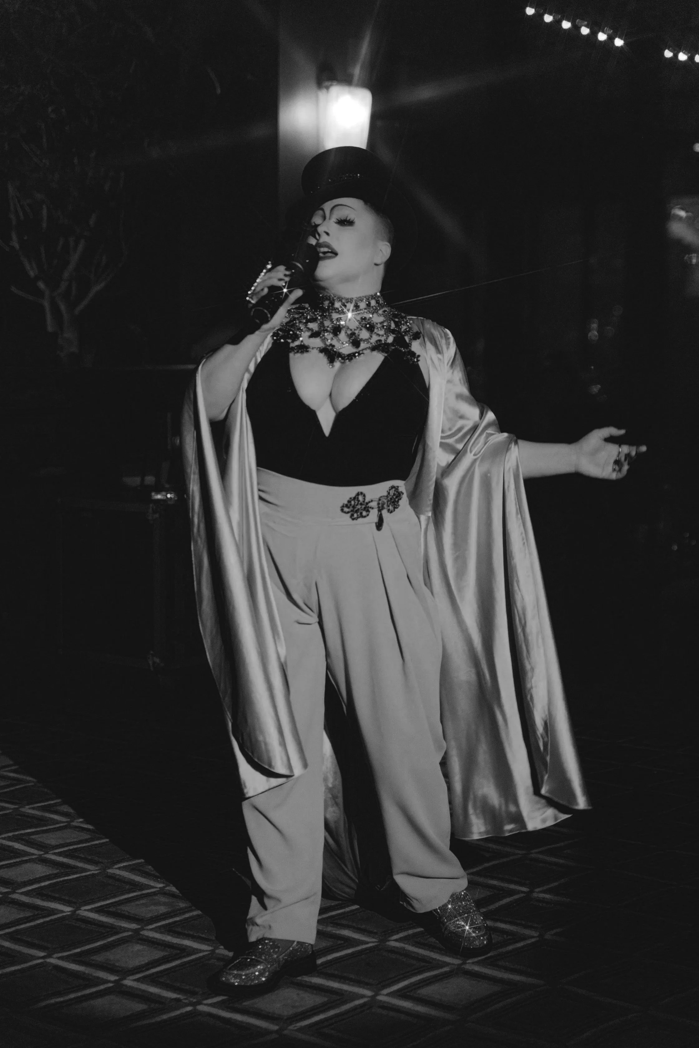 A performer singing into a microphone on stage, dressed in a shiny silk robe, wide-brimmed hat, and sparkly shoes, with elaborate makeup and jewelry.