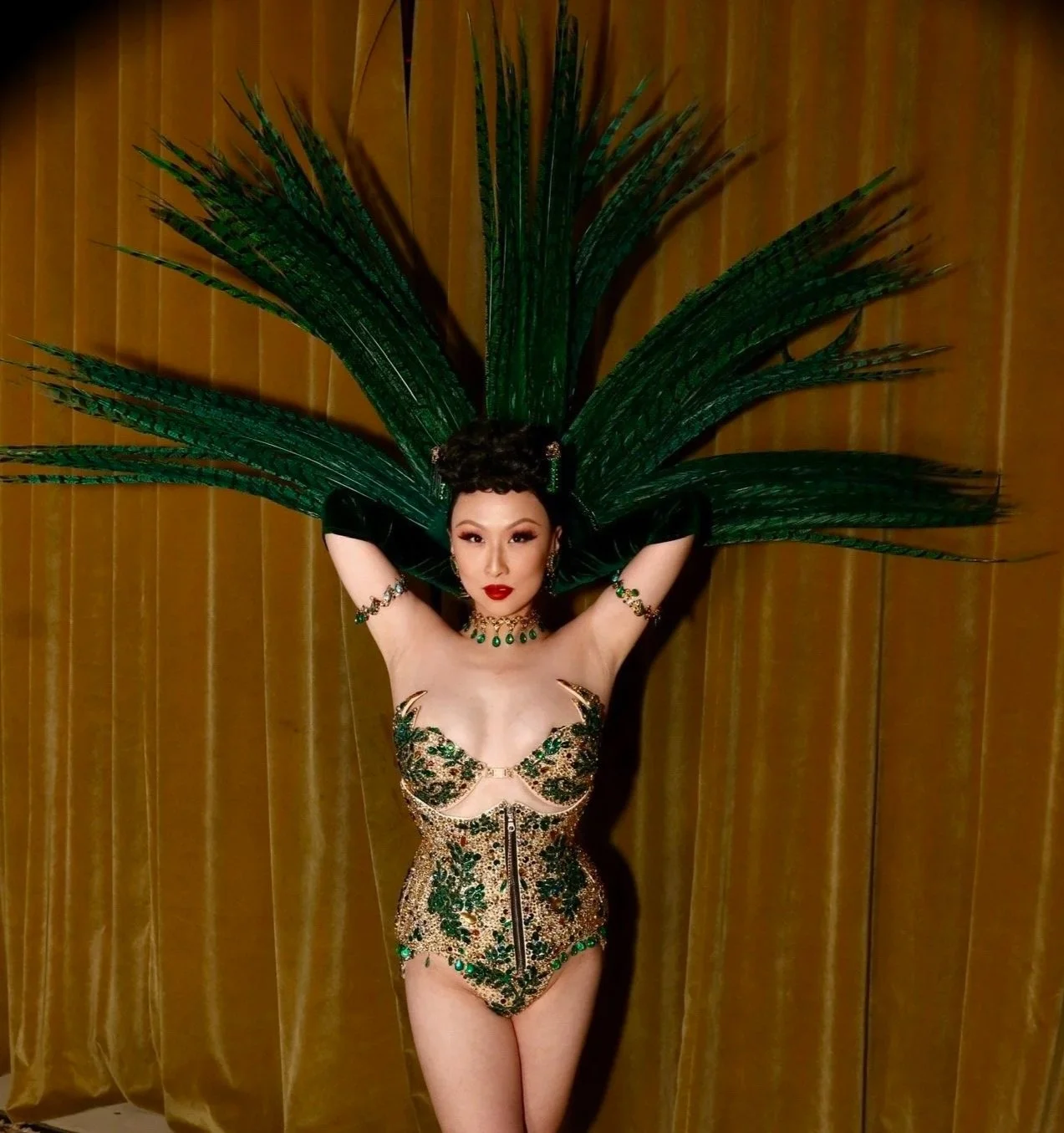A woman in an ornate costume with a large feathered headdress poses with arms raised against a gold curtain background.