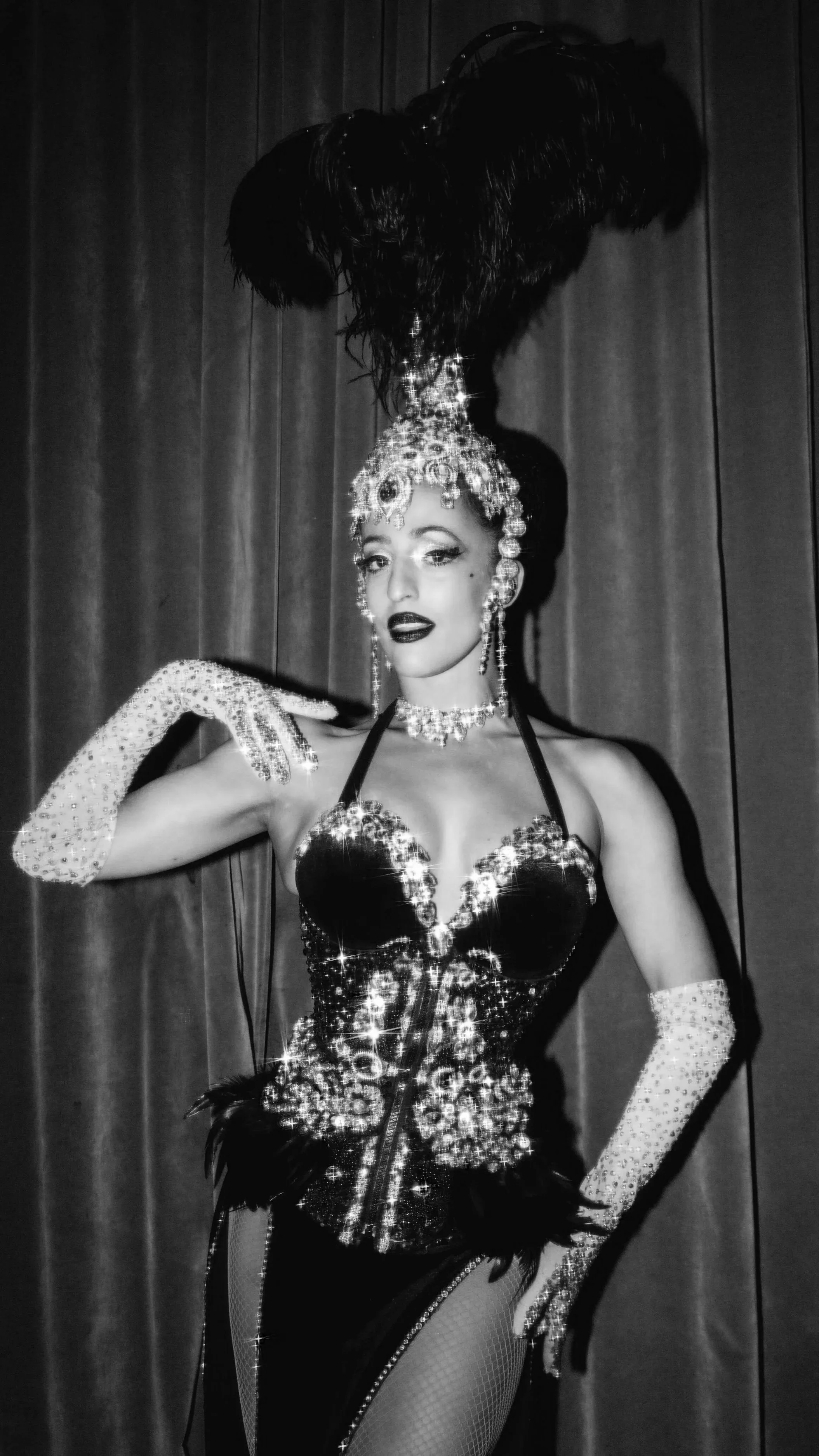 A woman in a glamorous, sparkling costume with a feathered headdress, jewelry, and long gloves, standing in front of a curtain.