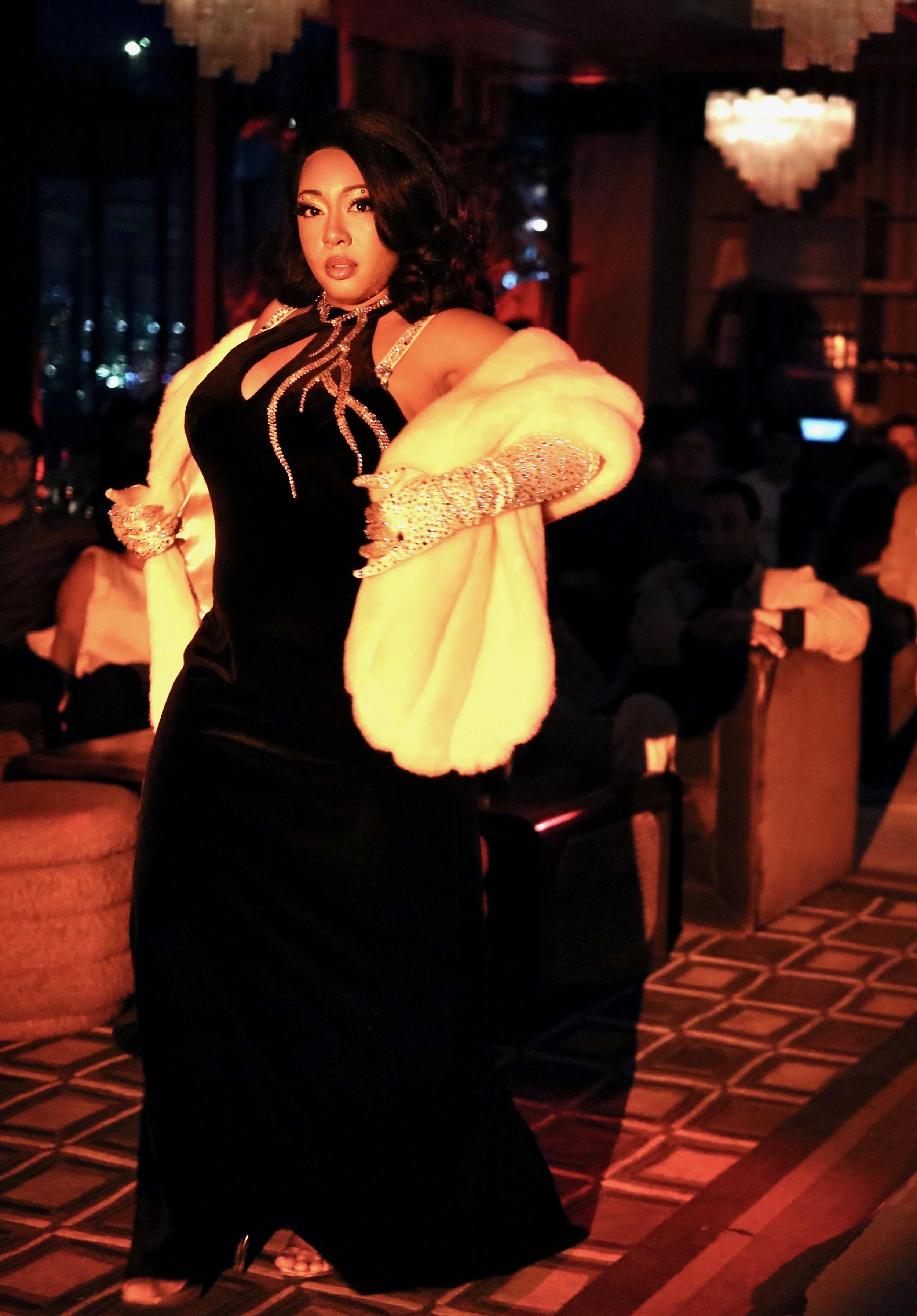 Woman in a black gown with rhinestone details, wearing a white fur stole and sparkling gloves, posing in a dimly lit, elegant setting with chandelier lighting and people seated in the background.