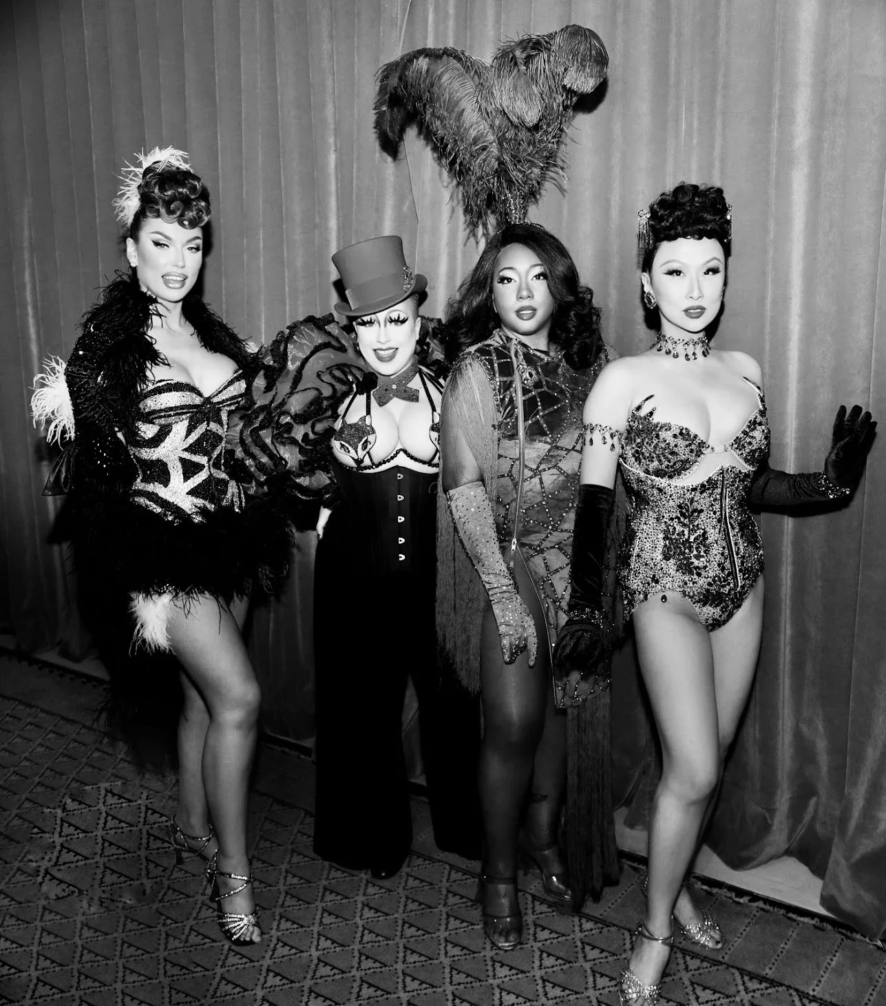 Four women dressed in showgirl costumes with feathers, sequins, and corsets, standing together in front of a curtain in a performance setting.