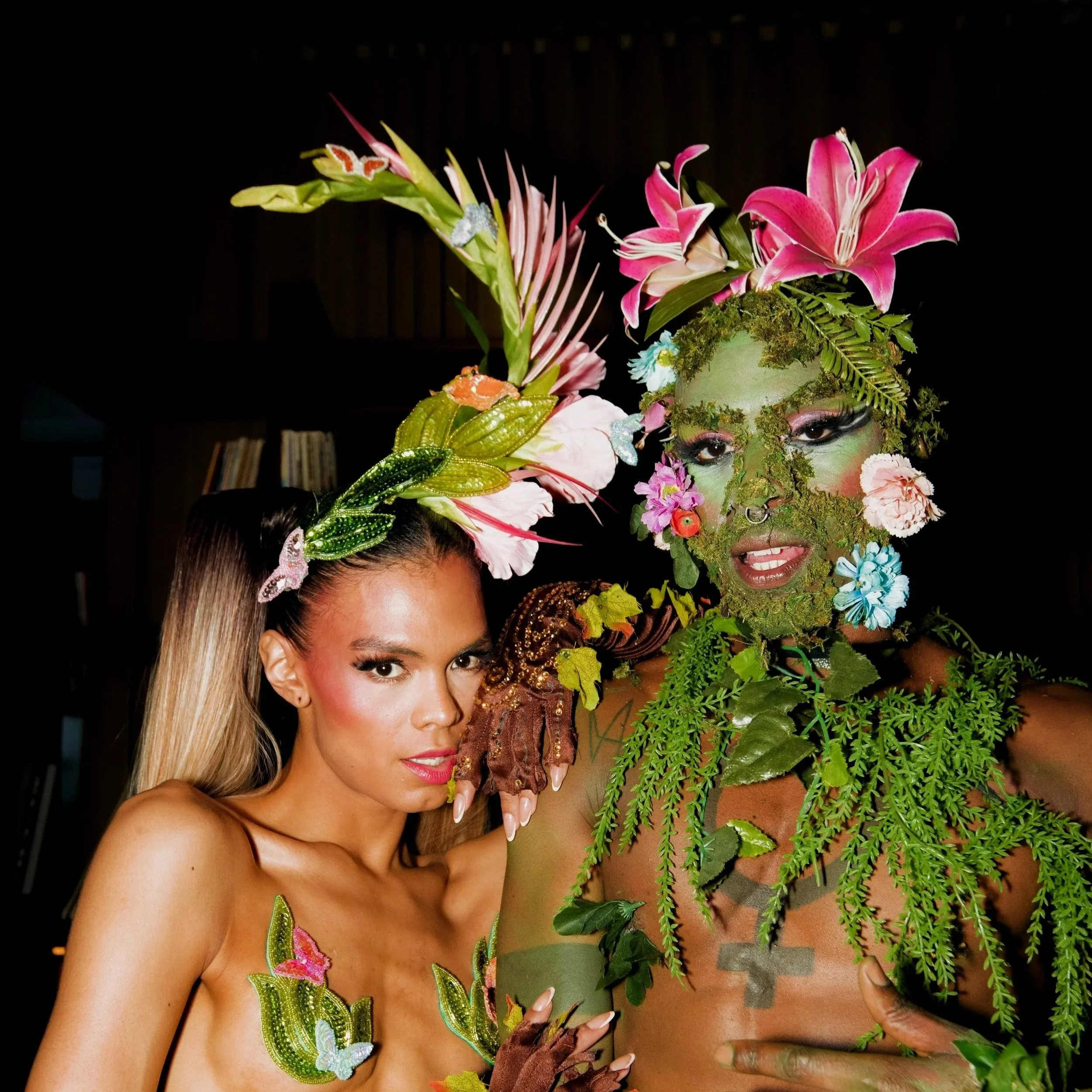 Two ambient performers dressed in elaborate costumes adorned with flowers and greenery, with one wearing headpieces made of large pink flowers and leaves, and the other face painted with moss and flowers, both posing together in a dark setting.