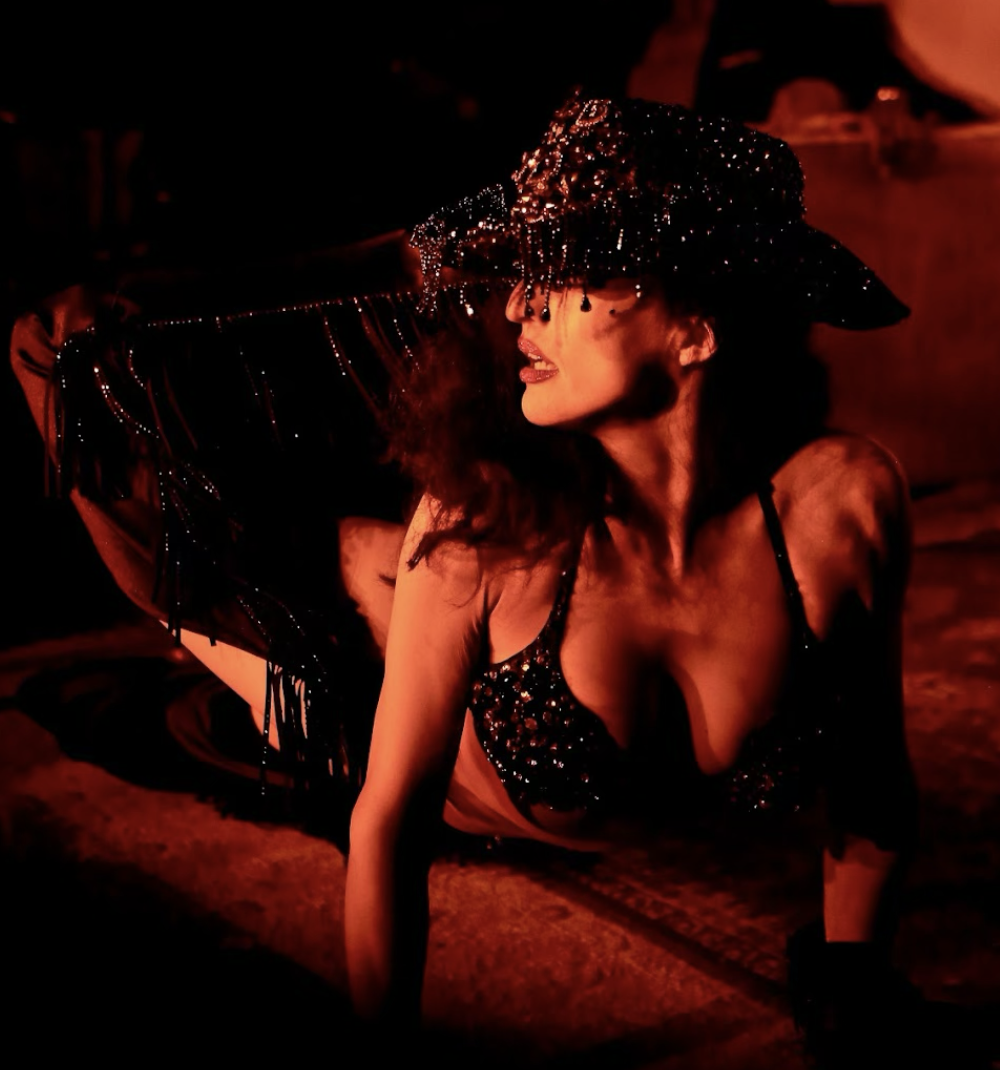 A performer wearing a black, sparkly, fringed dress and a wide-brimmed, sequined hat with hanging beads, posing on a darkly lit stage with red lighting.