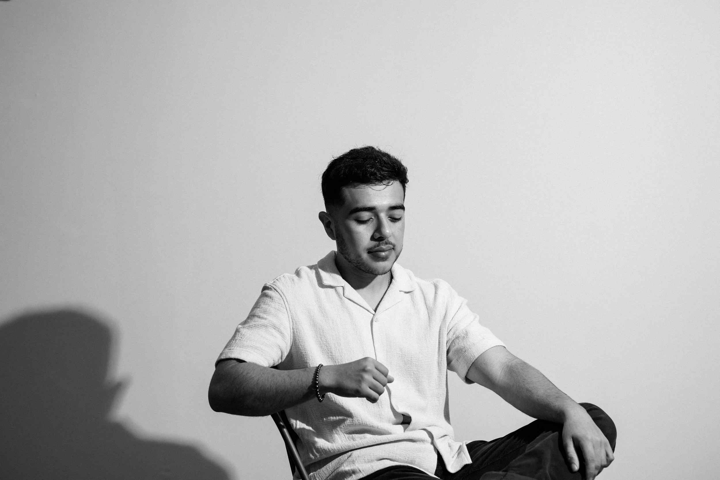 A young man with short dark hair and a beard sitting on a chair against a plain wall. He is wearing a light-colored, short-sleeved shirt and dark pants. He has a bracelet on his left wrist and appears to be looking down with a calm expression.