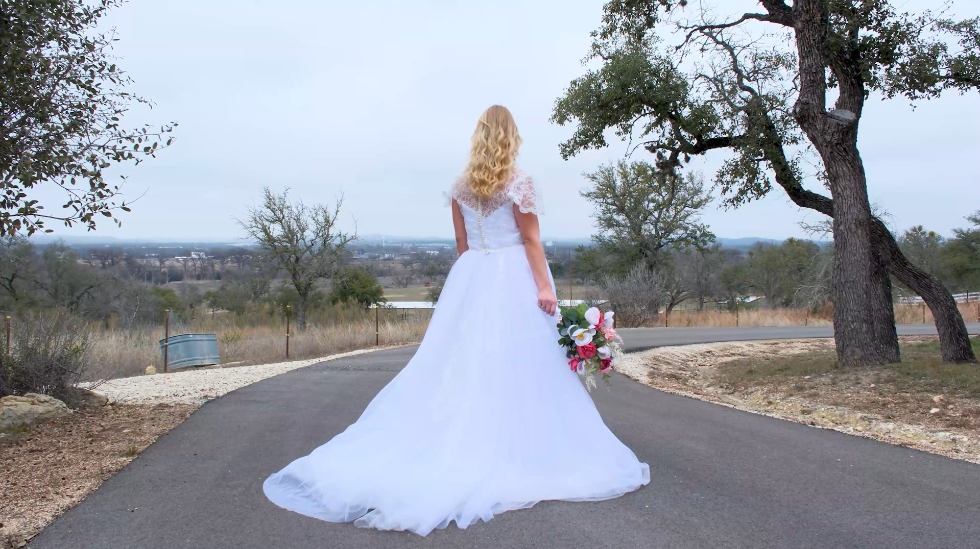 A bride in a white wedding gown holding a bouquet of pink and white flowers, standing on a rural road with trees and open landscape in the background.