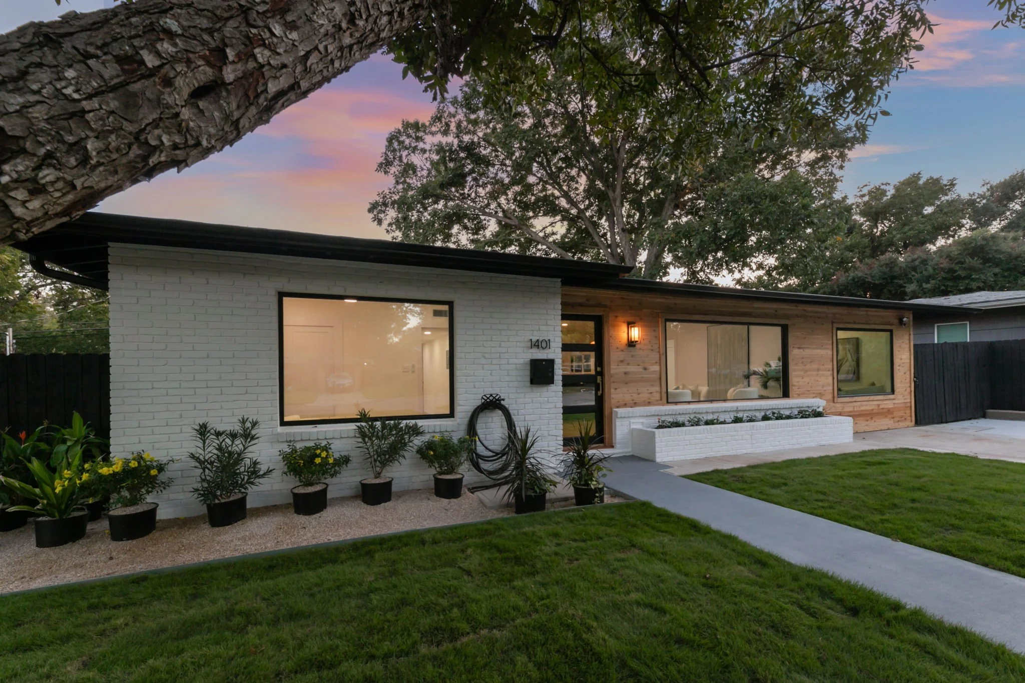 A modern single-story house with white brick and wooden exterior, large front windows, a garden with potted plants, and a green lawn at sunset.
