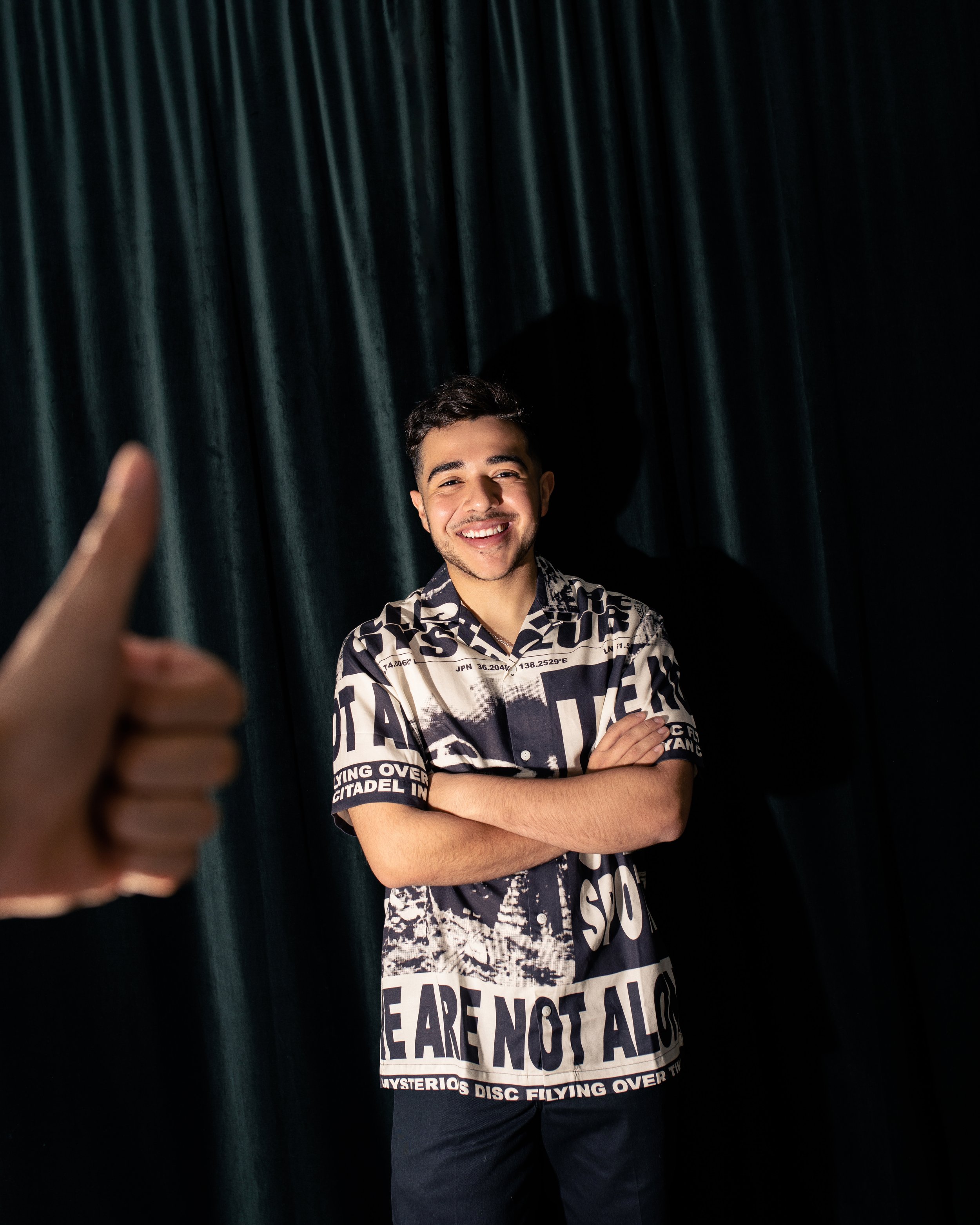 A young man with a big smile and crossed arms standing in front of a dark curtain. An out-of-focus hand giving a thumbs up is visible in the foreground.