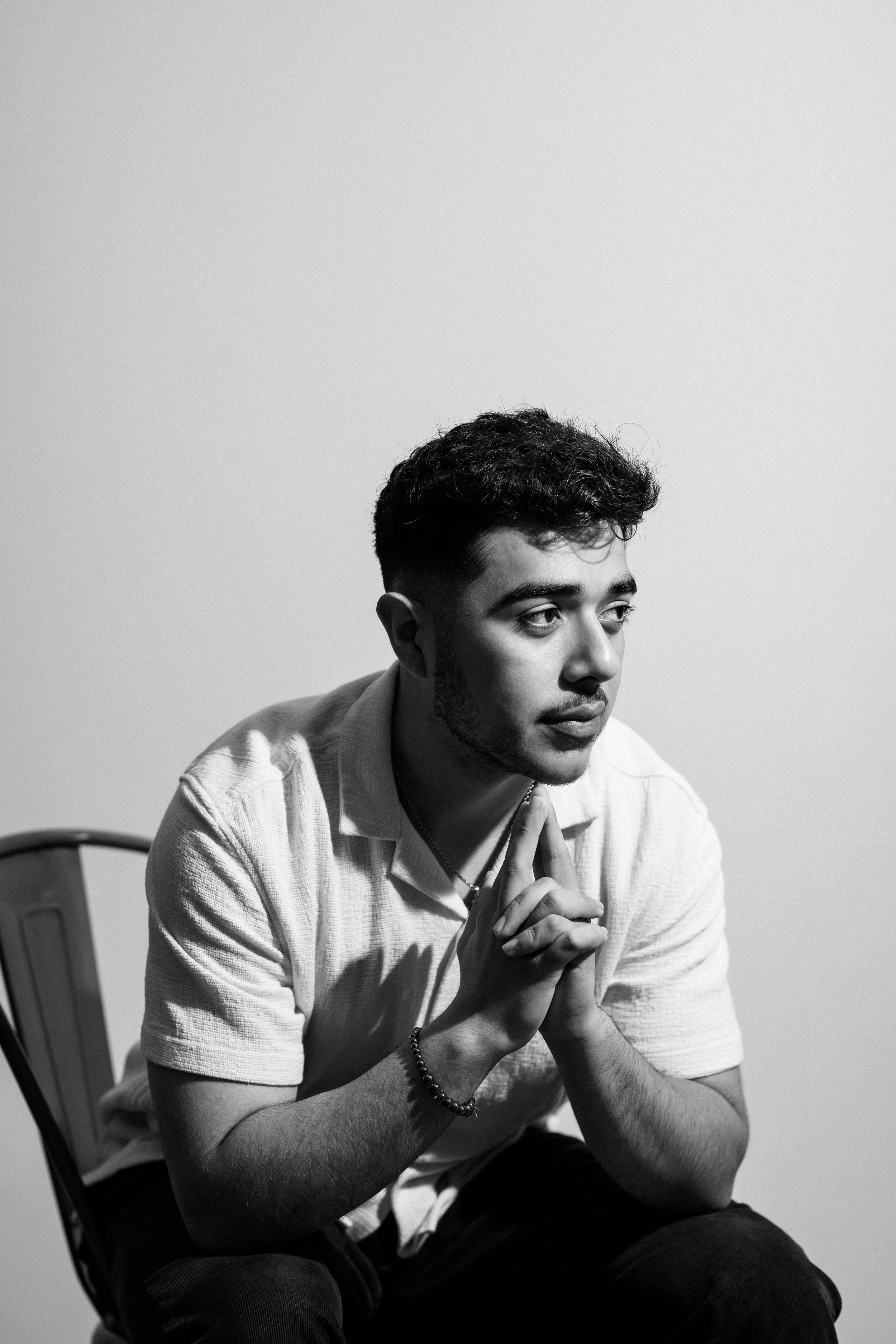 A young man with short curly hair and facial hair in a white polo shirt sitting on a chair with a plain background, clasping his hands together in front of his chin, looking contemplative in black and white.