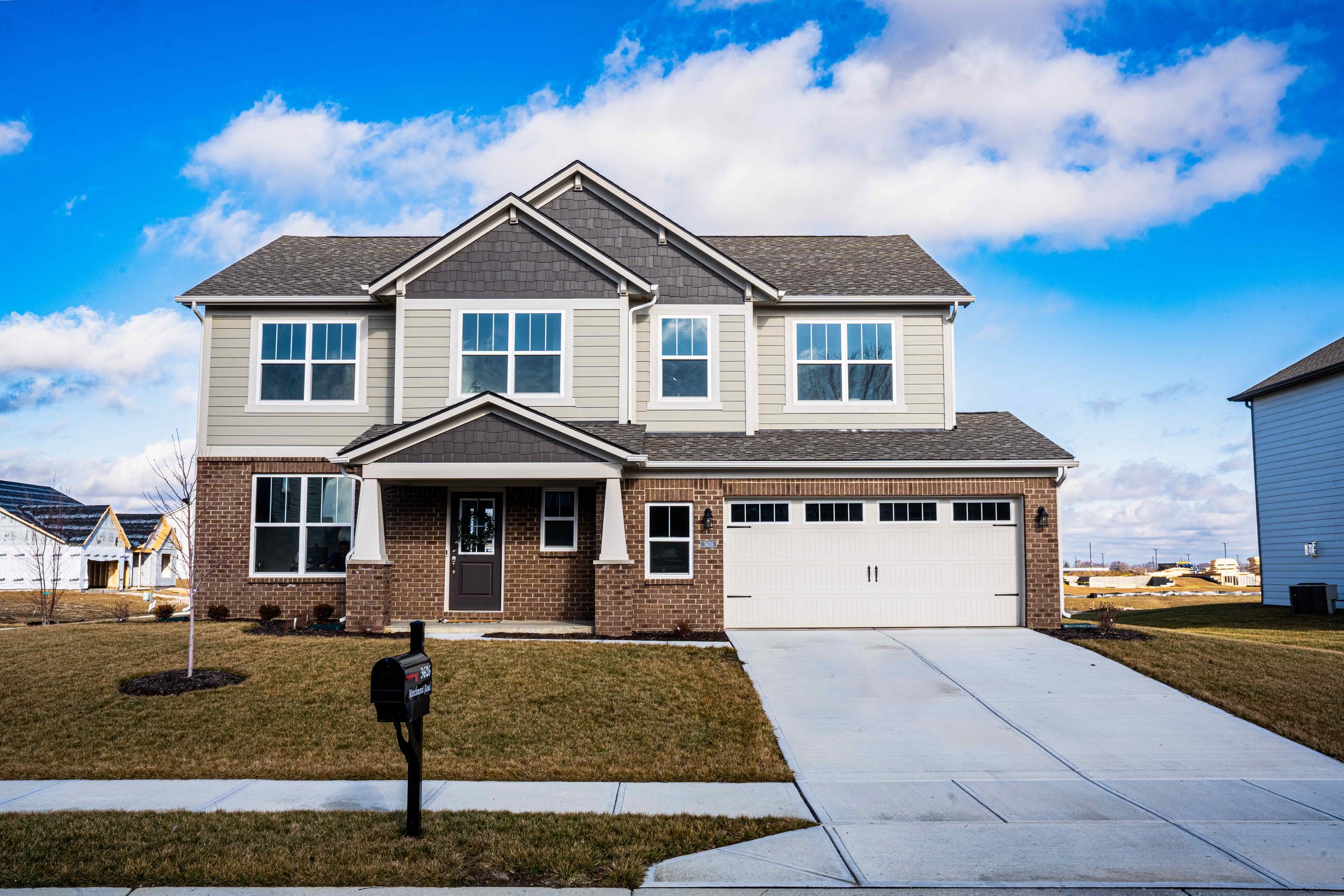 Newly built, two-story suburban house with grey siding and brick lower facade, attached garage, and a concrete driveway on a clear day with blue sky and clouds.